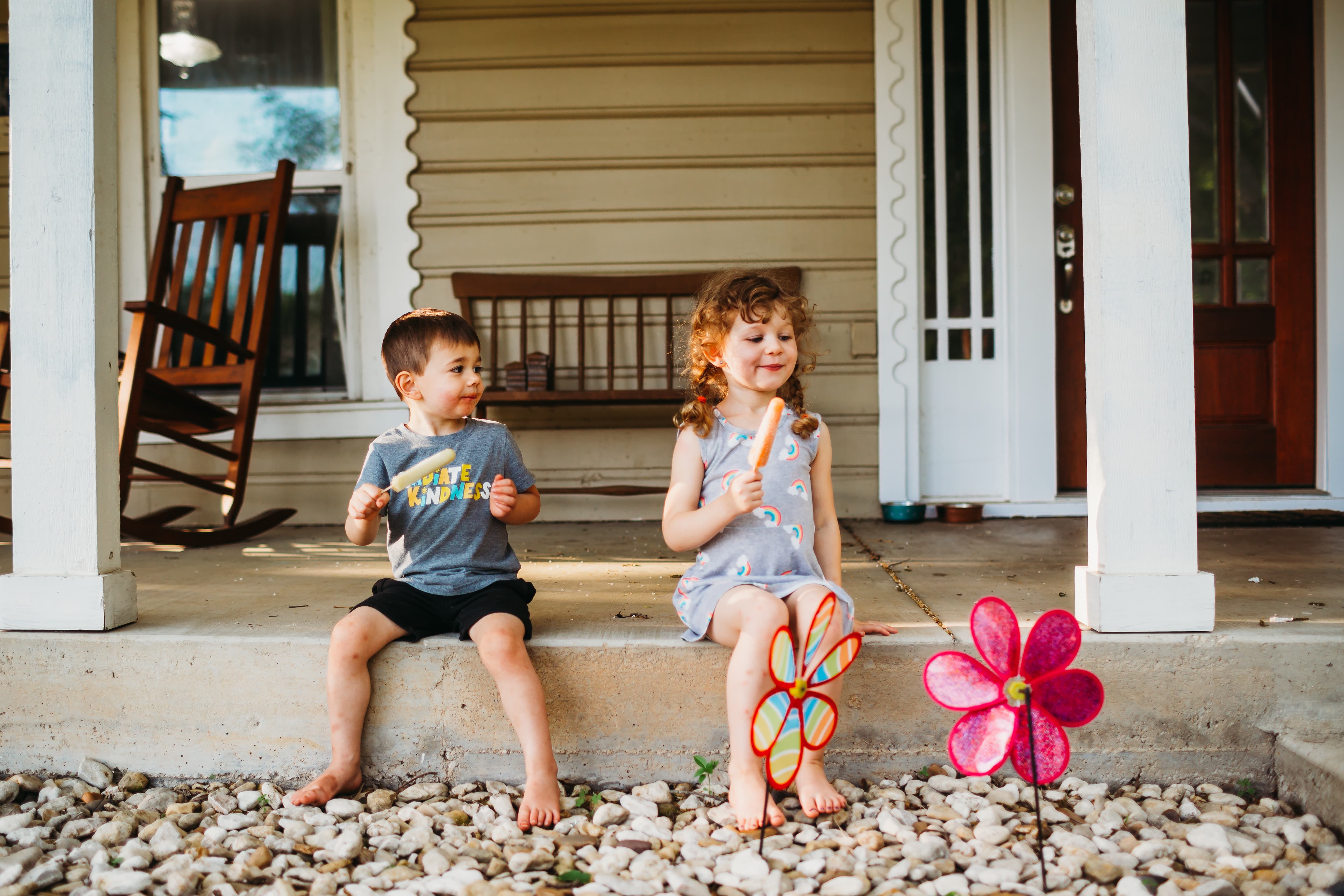 Brother and sister sitting on front porch eating popsicles in front of pinwheels
