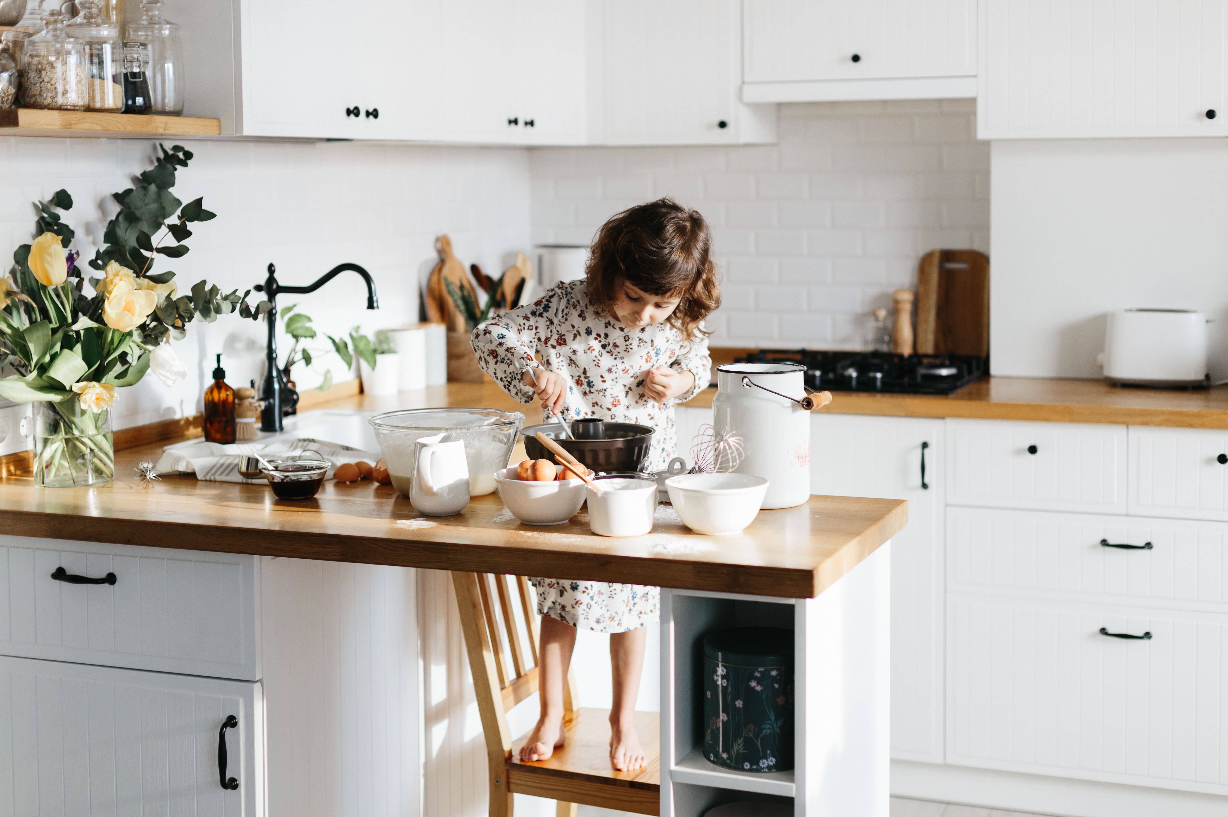A young girl stands on a stool to bake a cake in a small white kitchen