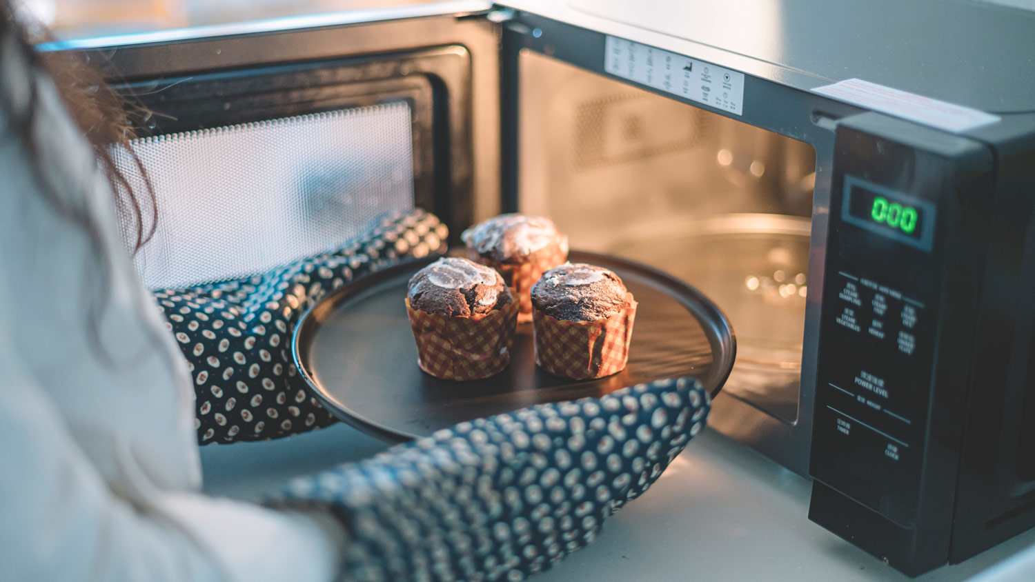 A woman wearing kitchen gloves heating cupcakes in microwave