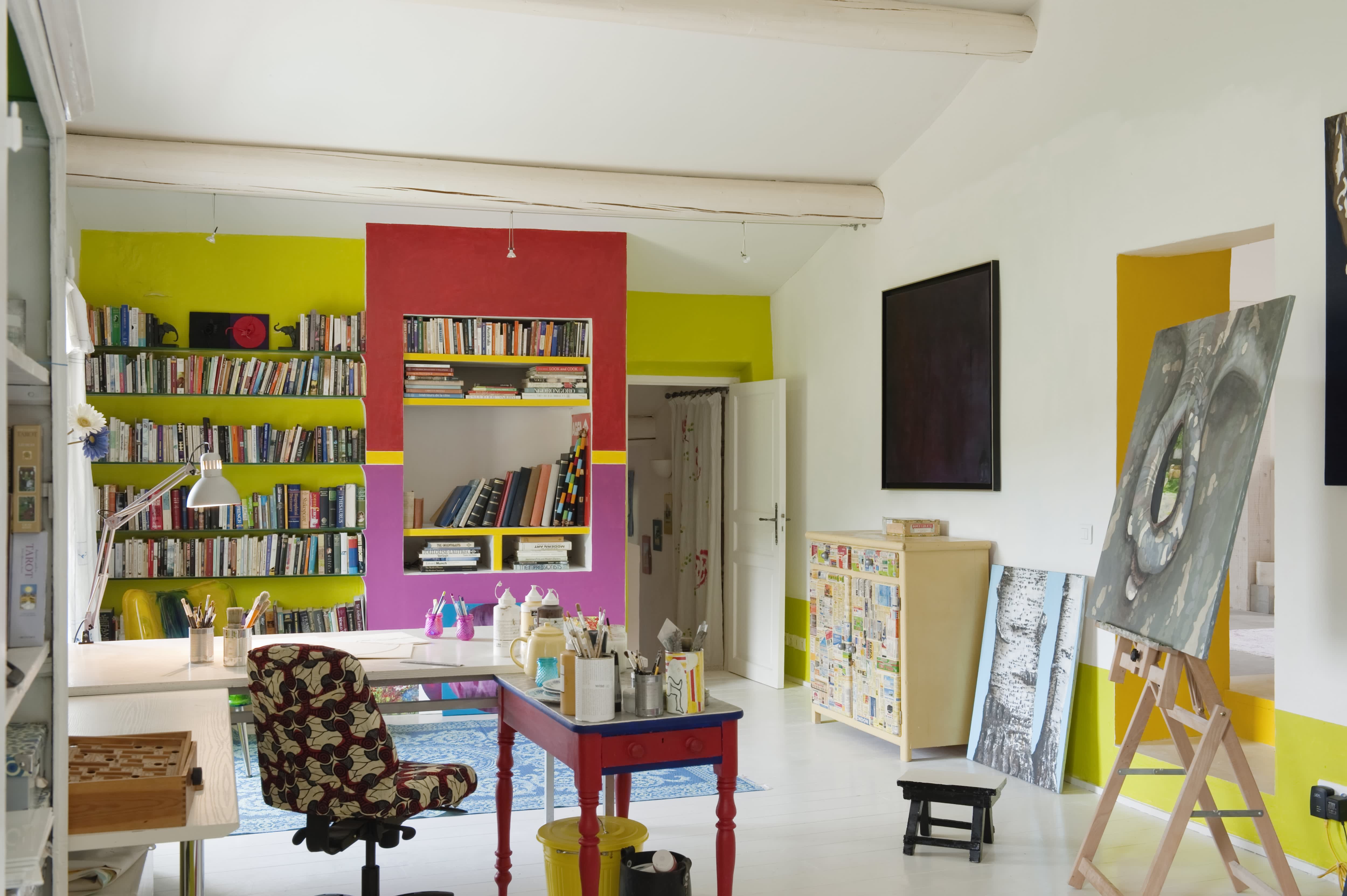 Bright, colorful craft room with built-in bookshelves, ceiling beams, a desk, paint supplies, and an easel