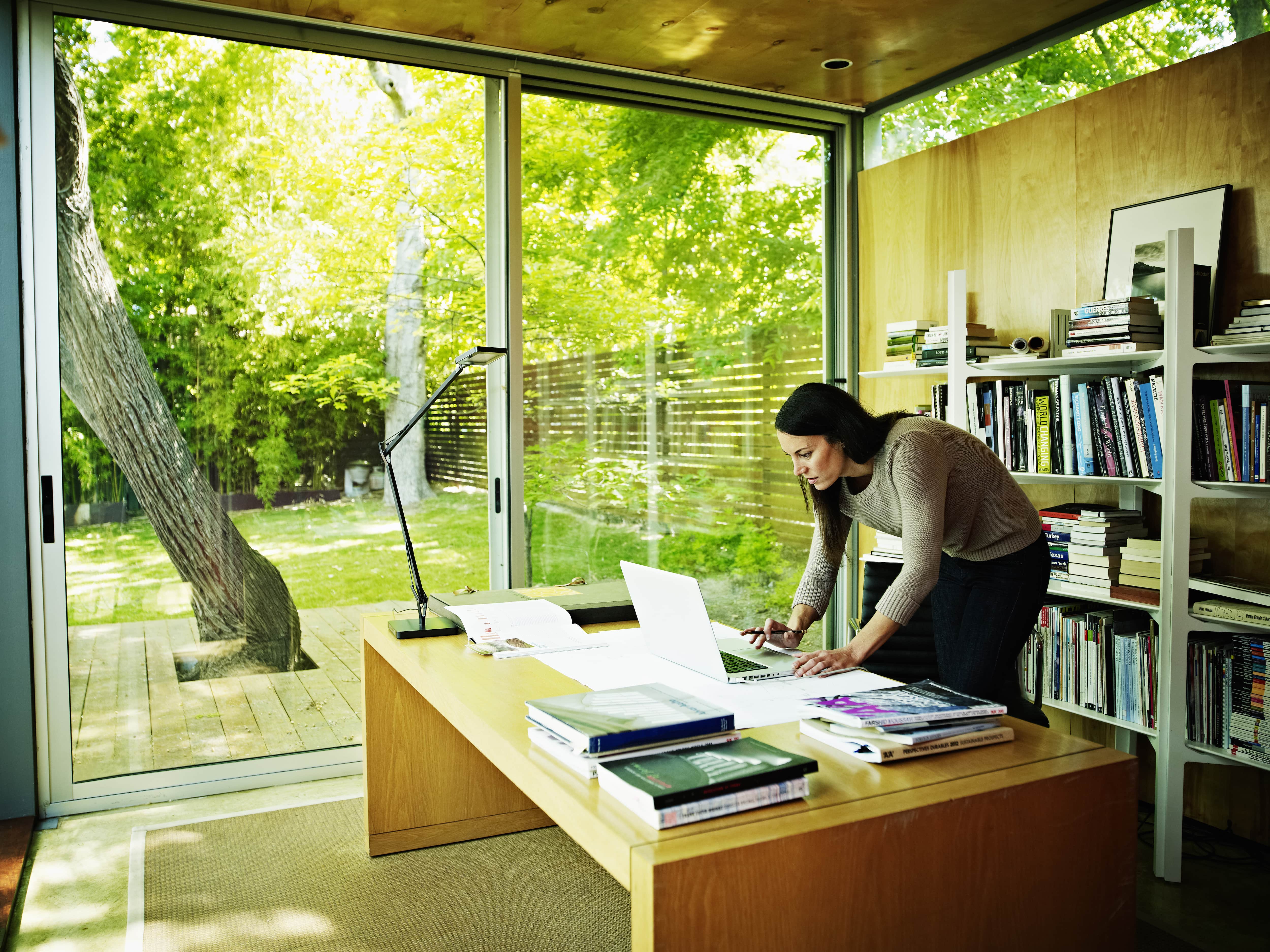 Woman working on laptop in home office with bookshelf and windows