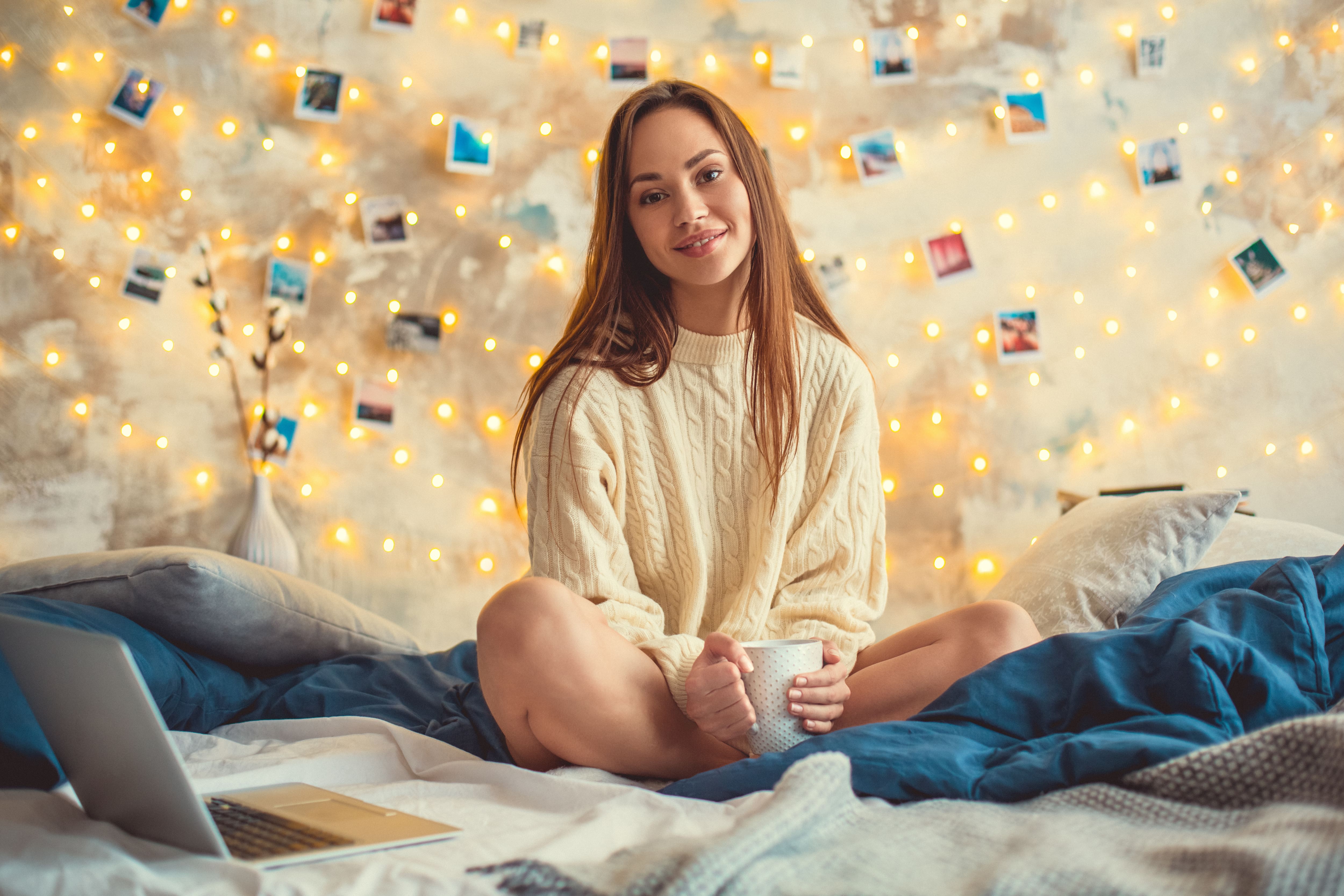 A young woman in a cozy sweater sitting on a bed in front of a string light wall