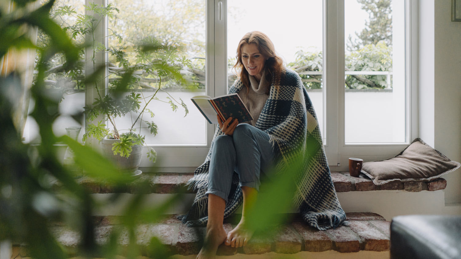 A woman wrapped in blanket reading a book on the sofa at home