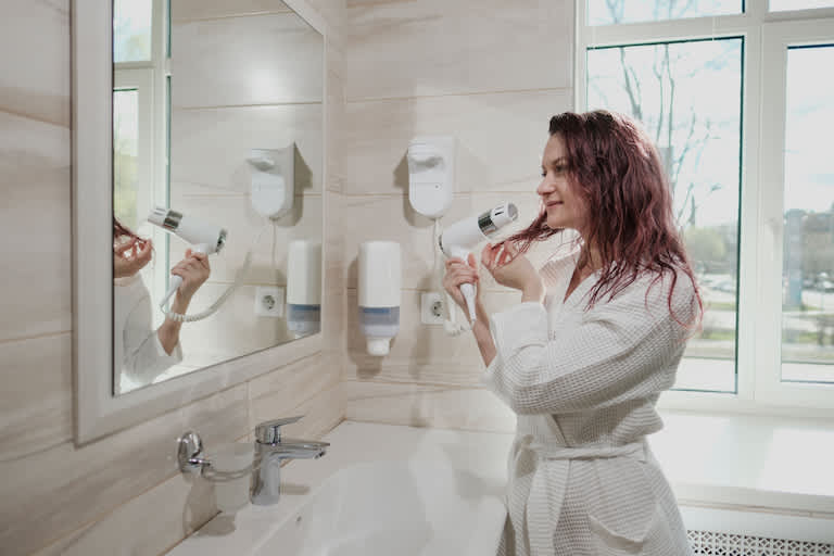 woman in white robe in white bathroom uses hairdryer and smiles