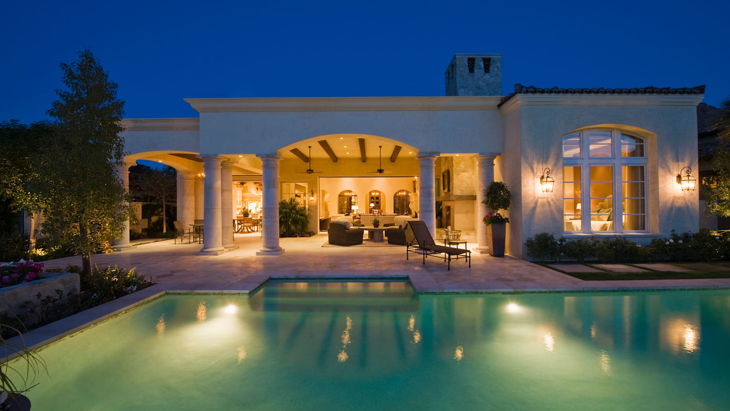 The exterior of a villa with lanterns on the walls and a lit swimming pool