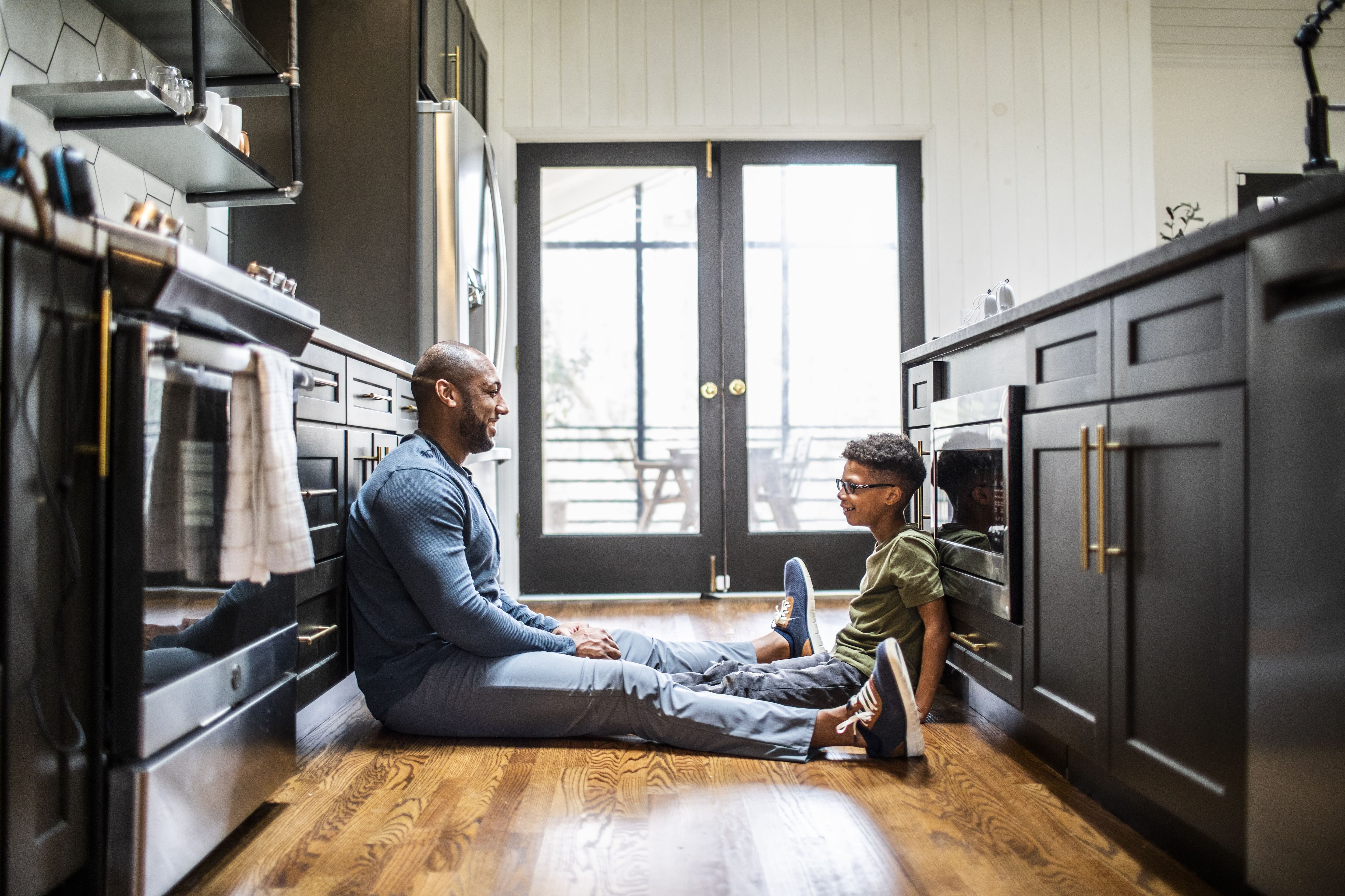 A father and son sitting on the floor of a galley kitchen while talking
