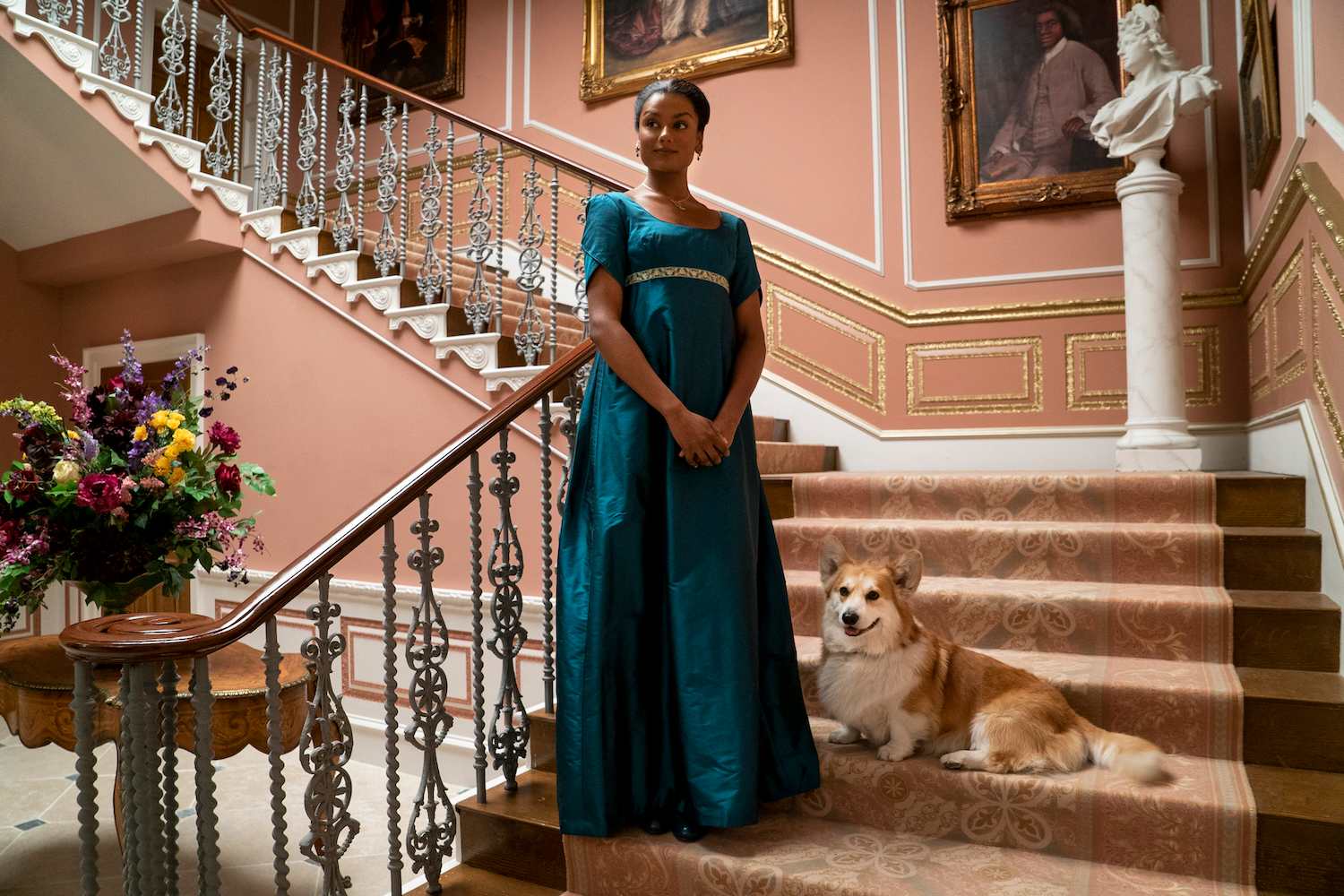 Woman in blue Regency dress standing on ornate staircase with a Corgi sitting beside her