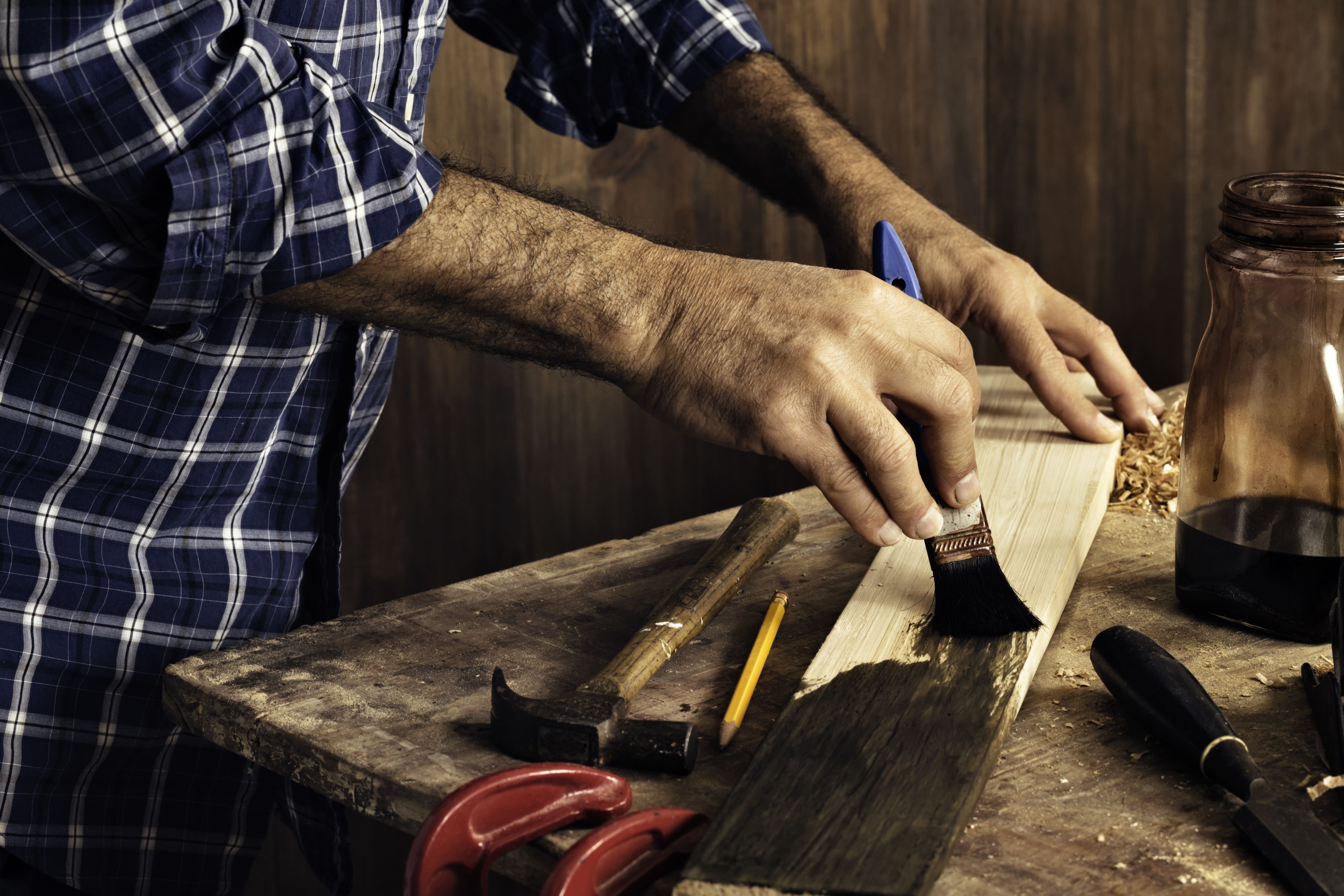 A male carpenter in a plaid shirt staining a wood plank