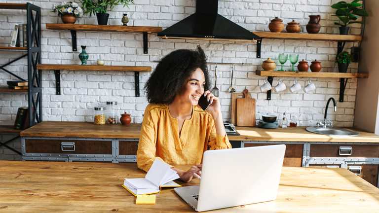 Black woman in her kitchen talking on phone at laptop
