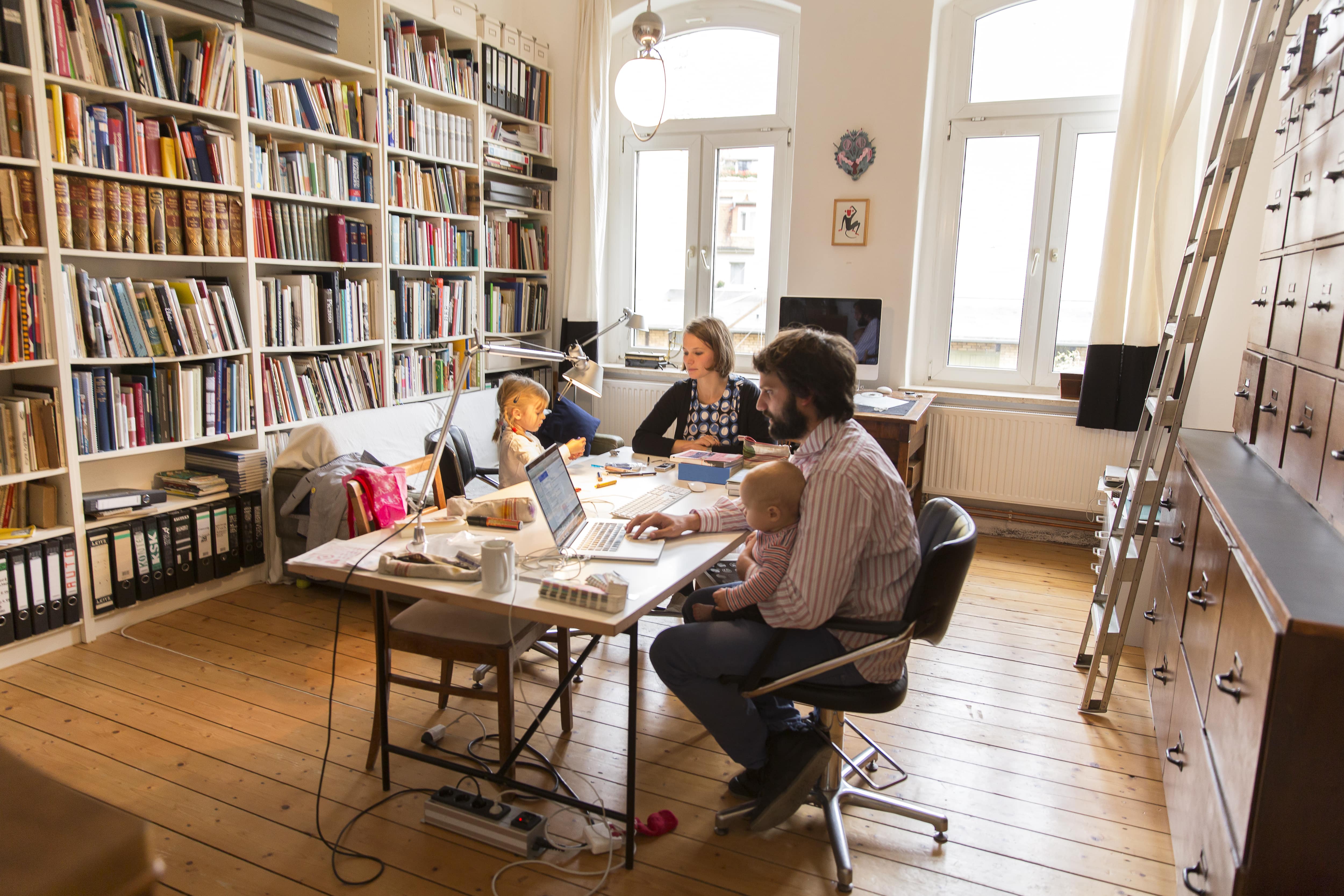 Husband and wife designers work at a shared desk in their home library along with their children