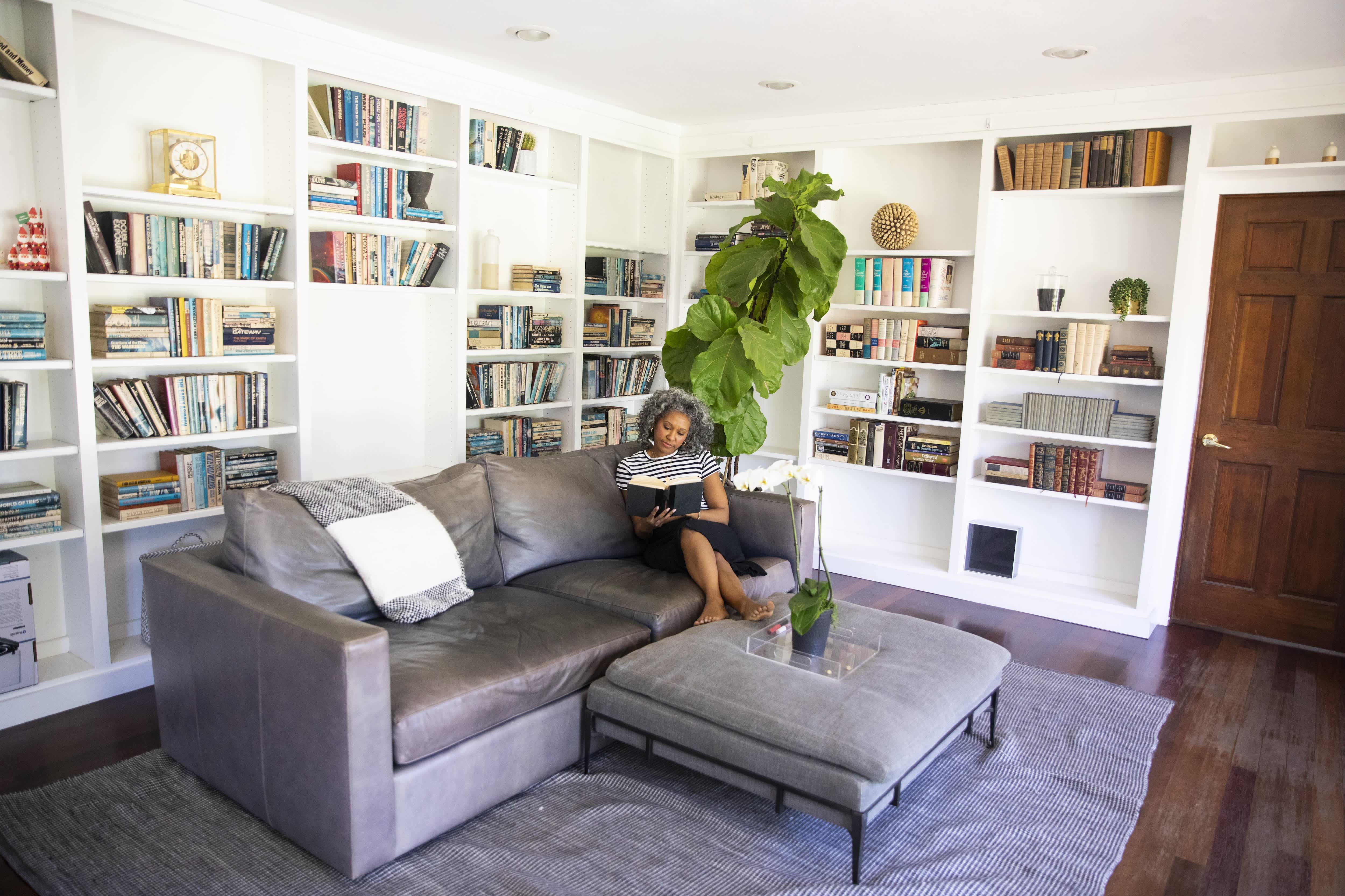 Black woman with curly white hair reading a book on a gray leather couch in her home library