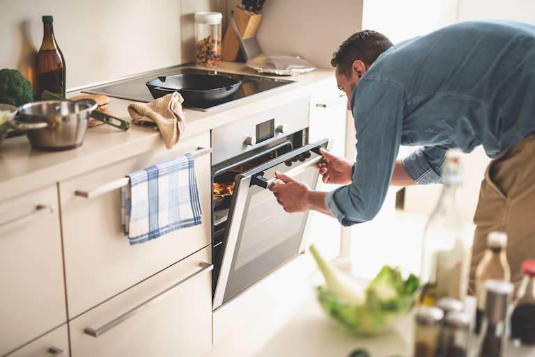 man opening oven to check on food inside in bright, cream colored kitchen strewn with various cooking tools