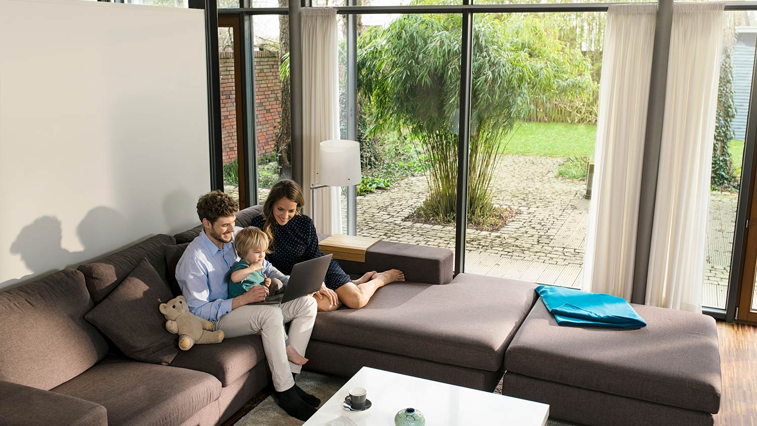 Parents and son on sofa in living room with large windows