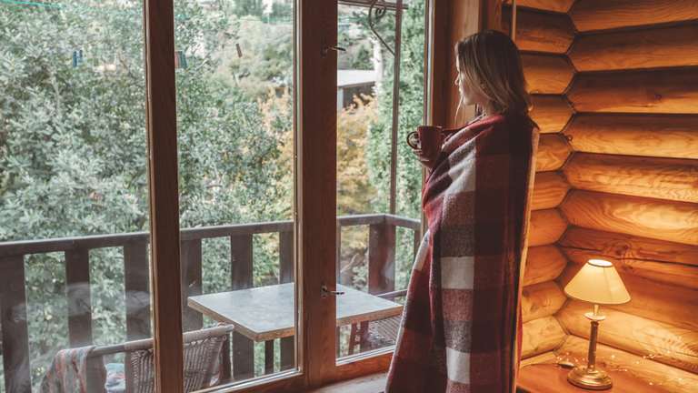 Woman wrapped in blanket looking outside of log house