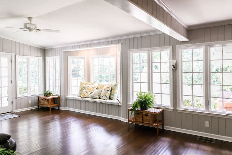 Large and open living room leading into sunroom with windows on two sides and lots of natural light flowing in. There is a window seat on one side and a leather couch and plant on the other.