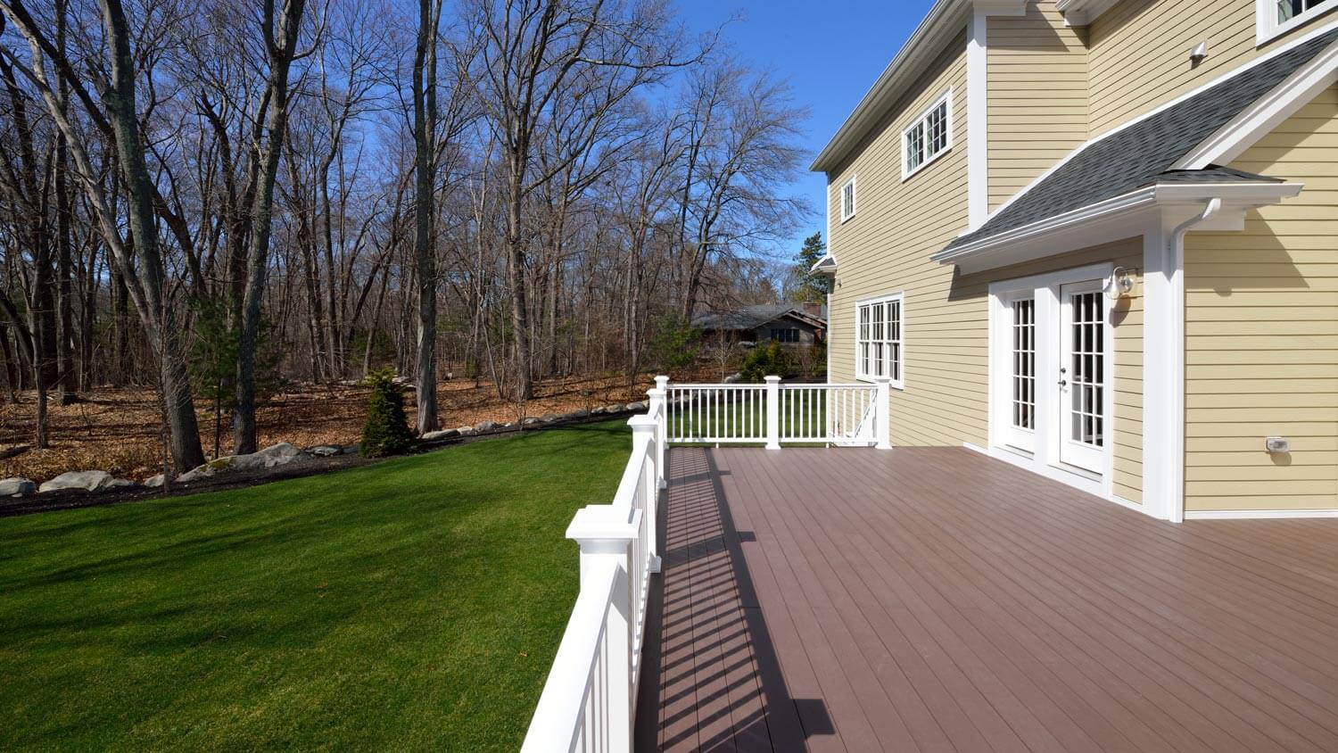 light tan deck with green grass and bushes surround it and silver wire deck chairs and table on top