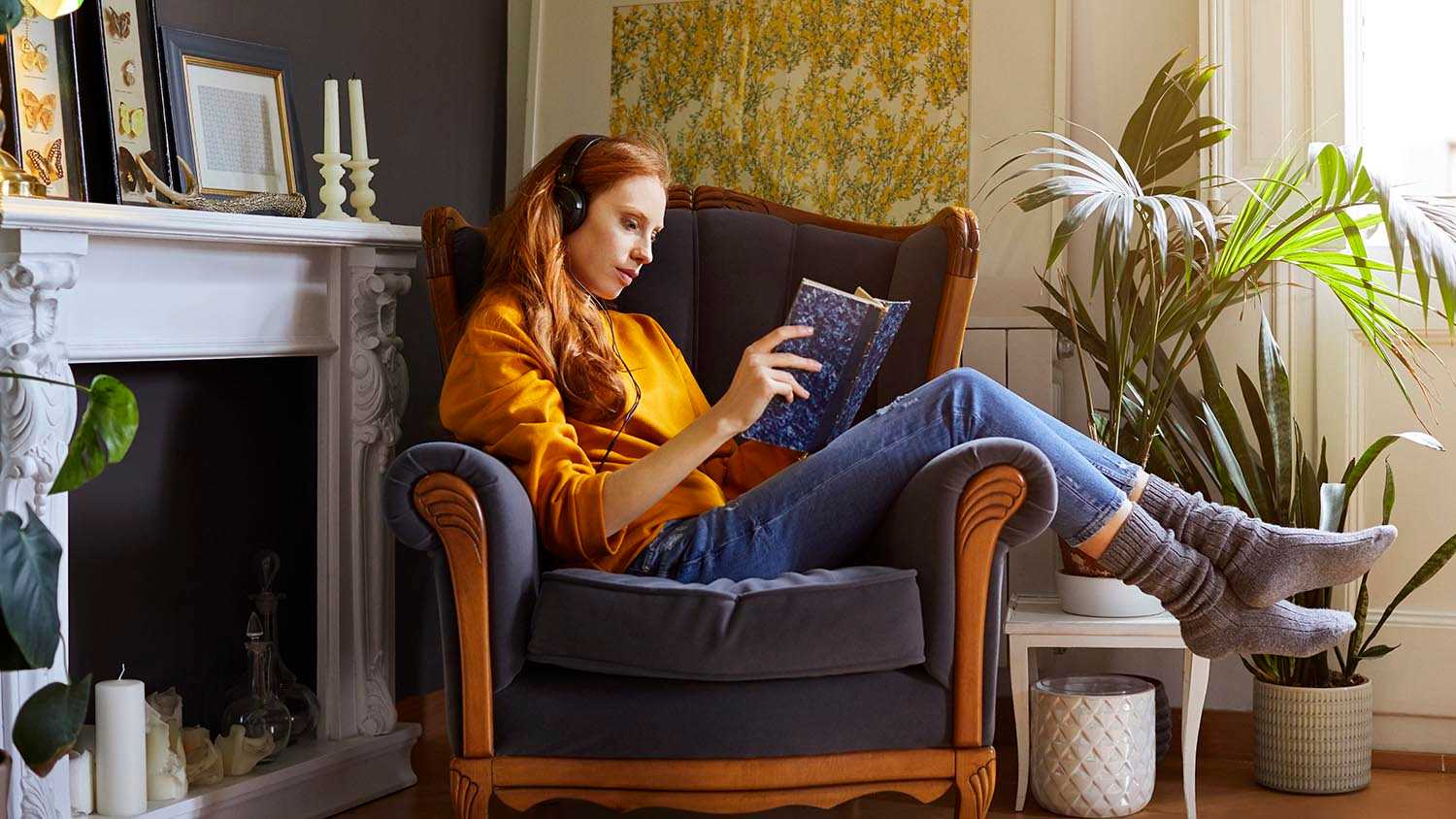woman with headphones on reading on chair next to a fireplace