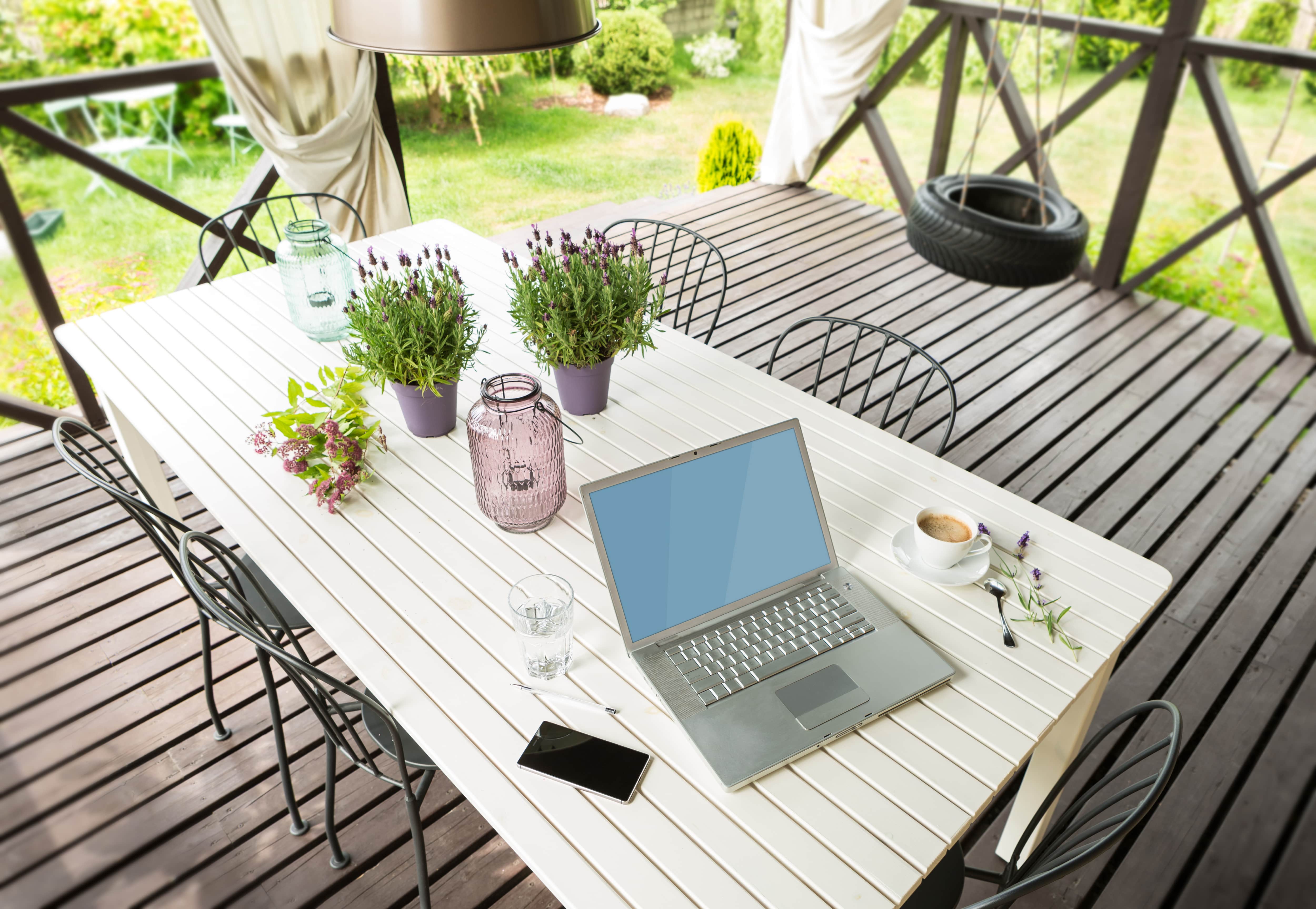A porch with a large white table with a laptop, latte, phone, and potted plants on top