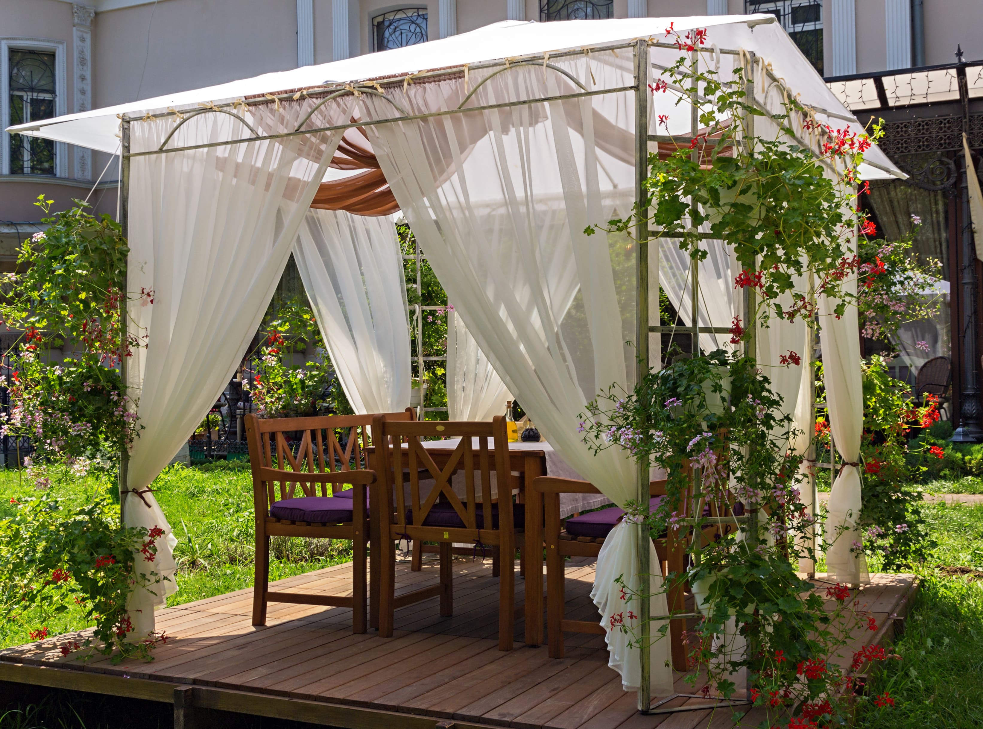 Pergola with a white roof and white sheer curtains surrounded by flowers