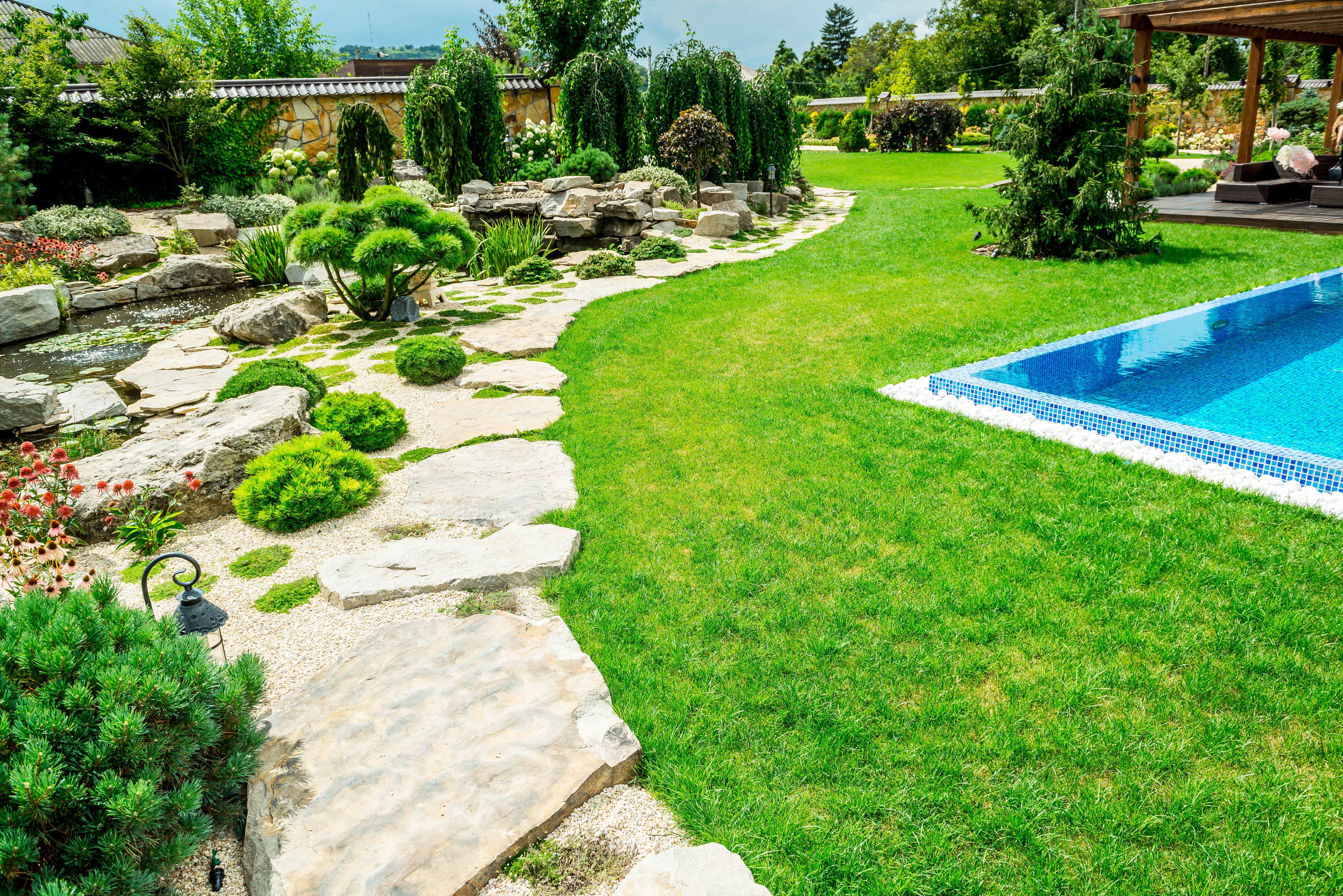 A backyard in-ground pool surrounded by grass with outer perimeter solid rock hardscaping