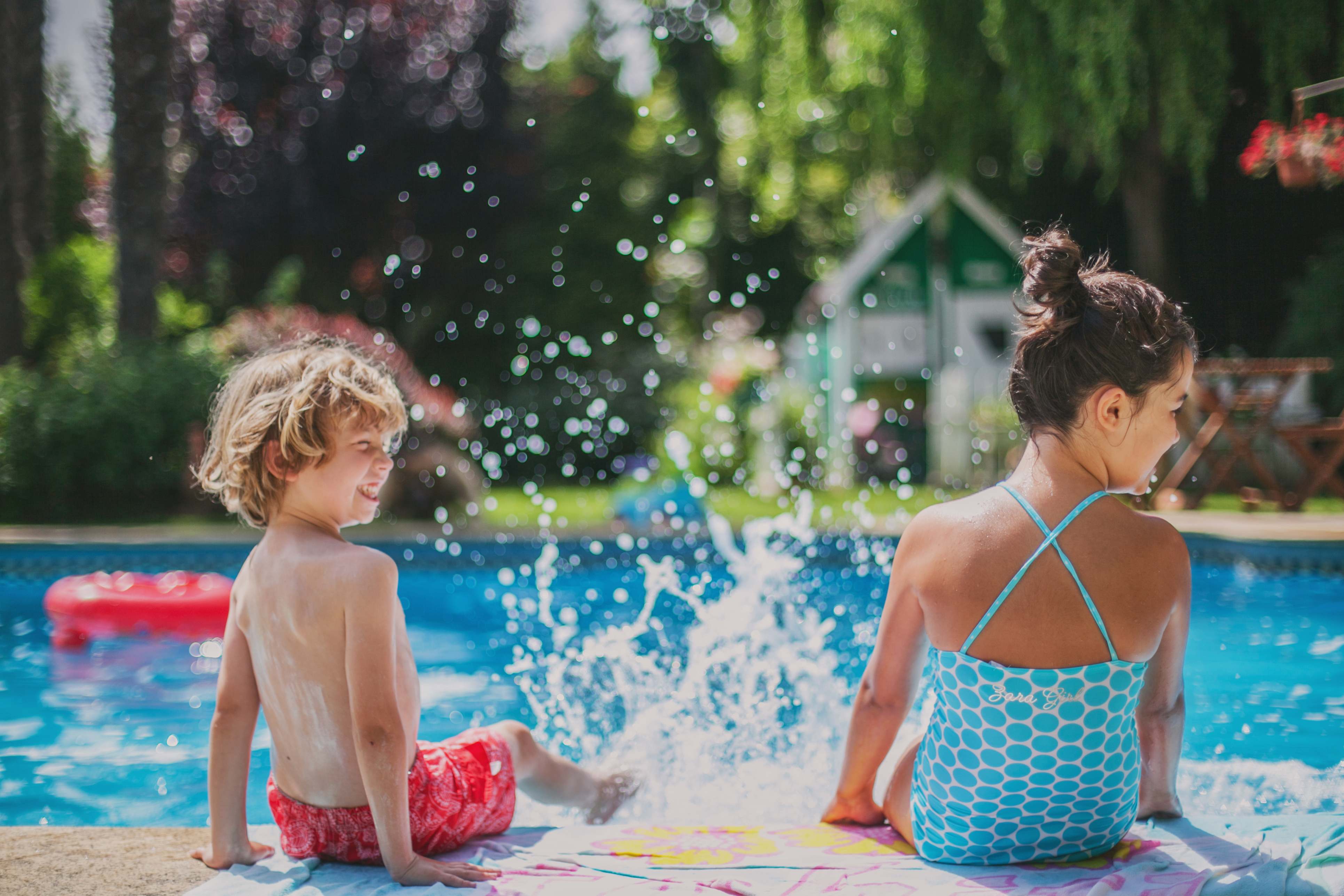 Two kids kicking water in an inground pool next to a small green pool house