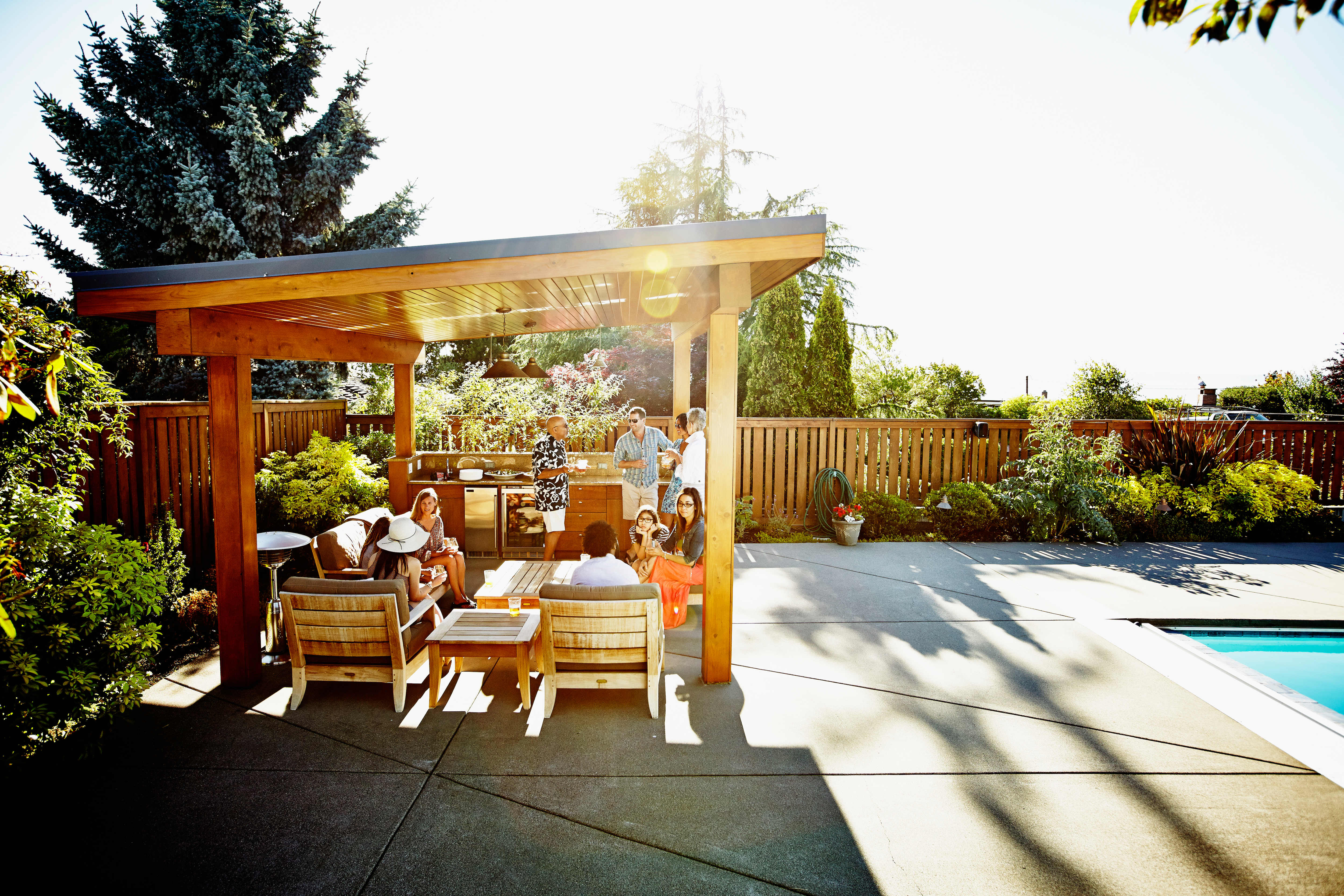 A group of people enjoying a cookout under a pergola next to an inground pool