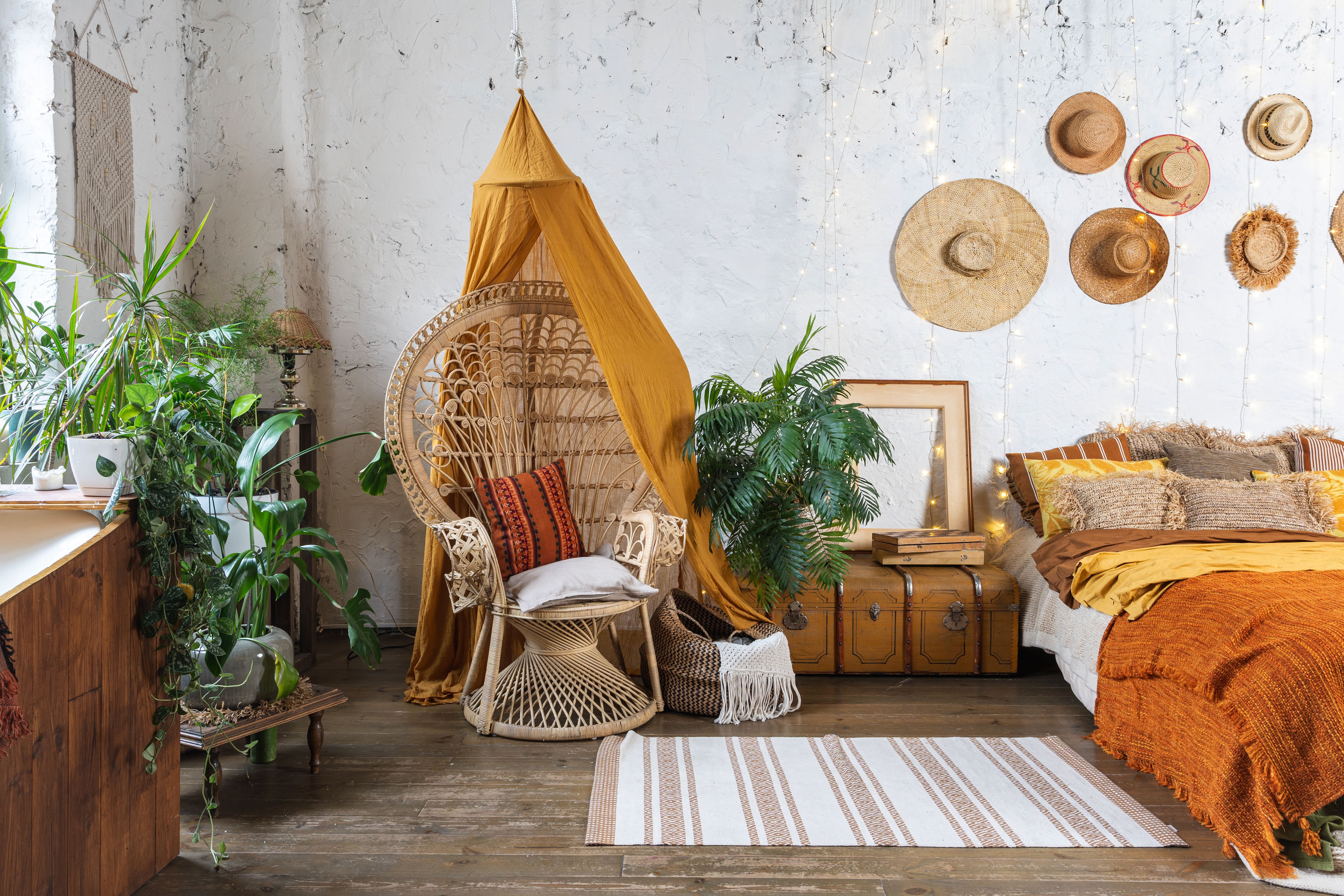 Boho bedroom with orange canopy over rattan chair, vintage suitcase, plants, and hats and fairy lights on the wall