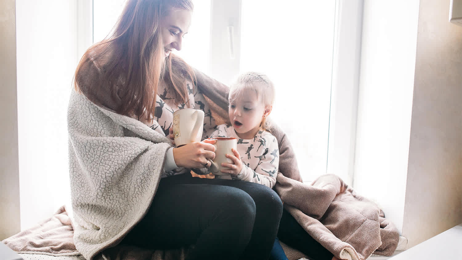 Mom and daughter are cozy under blanket with hot chocolate