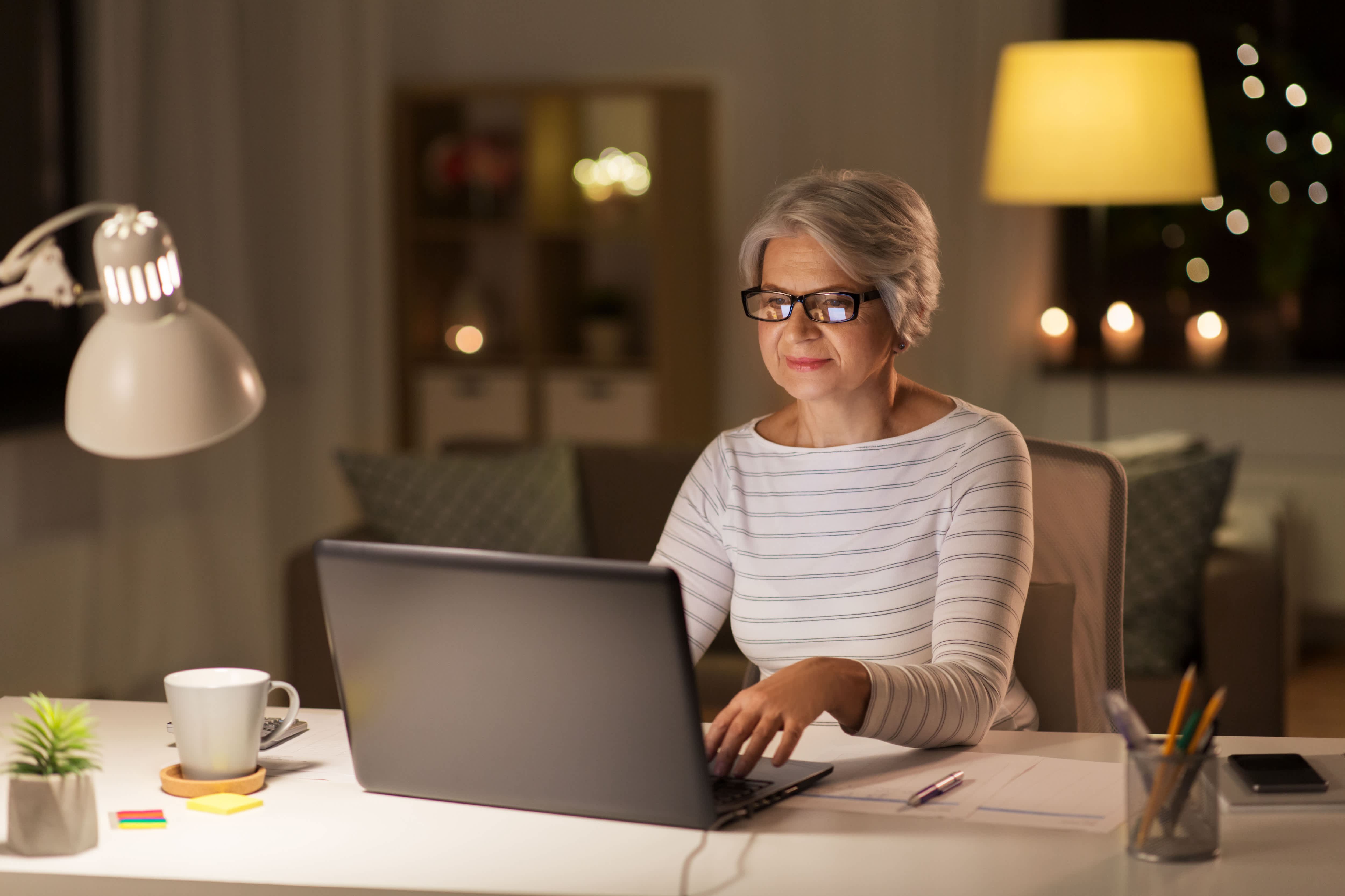 Older woman in glasses working on her laptop in a softly lit home office