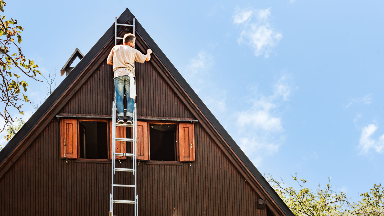 Man on Ladder Protecting Wooden House Exterior Applying Paint with Paintbrush