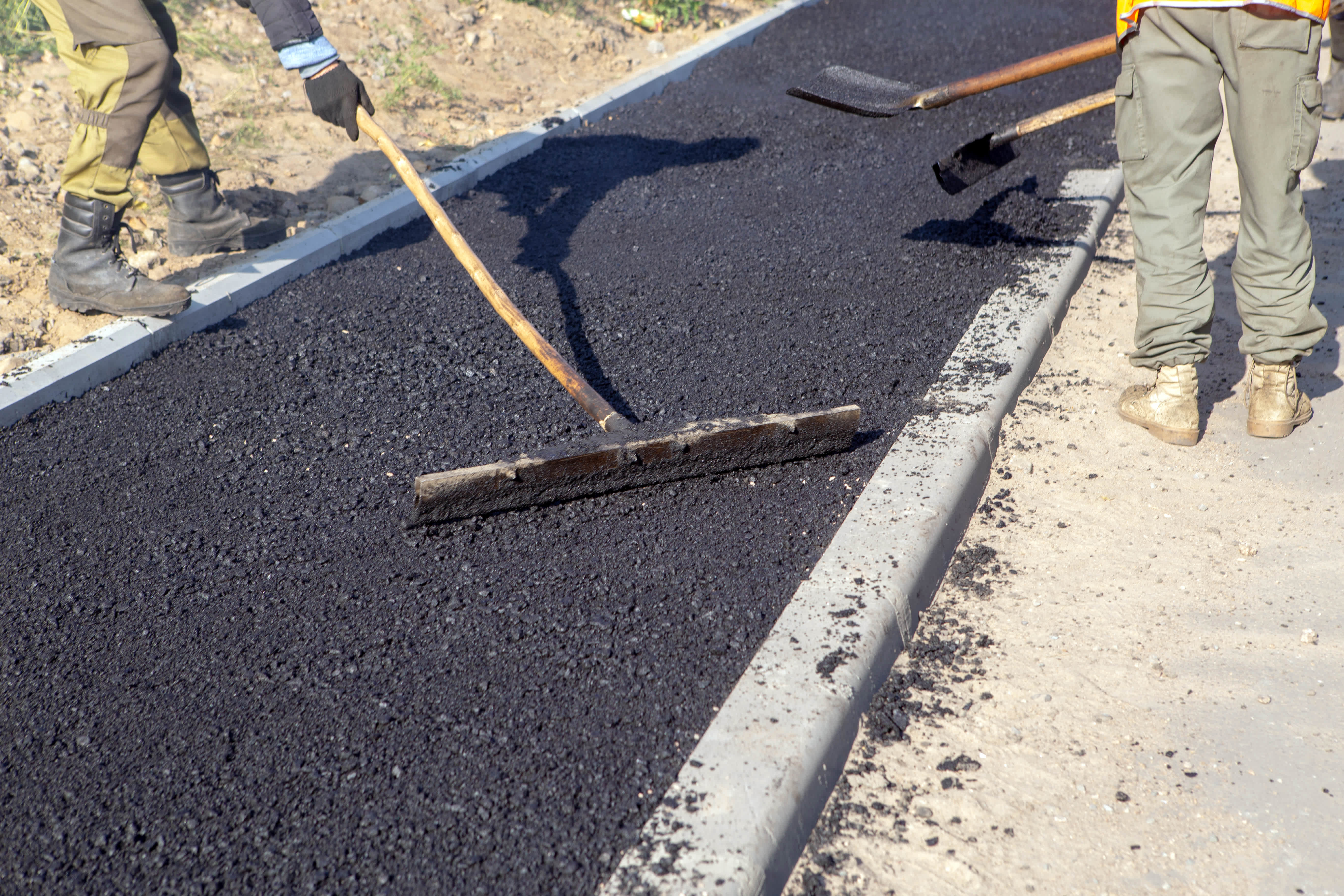 Workers use a squeegee tool to spread and level hot asphalt during an asphalt install, ensuring a smooth and even surface for the new pavement.