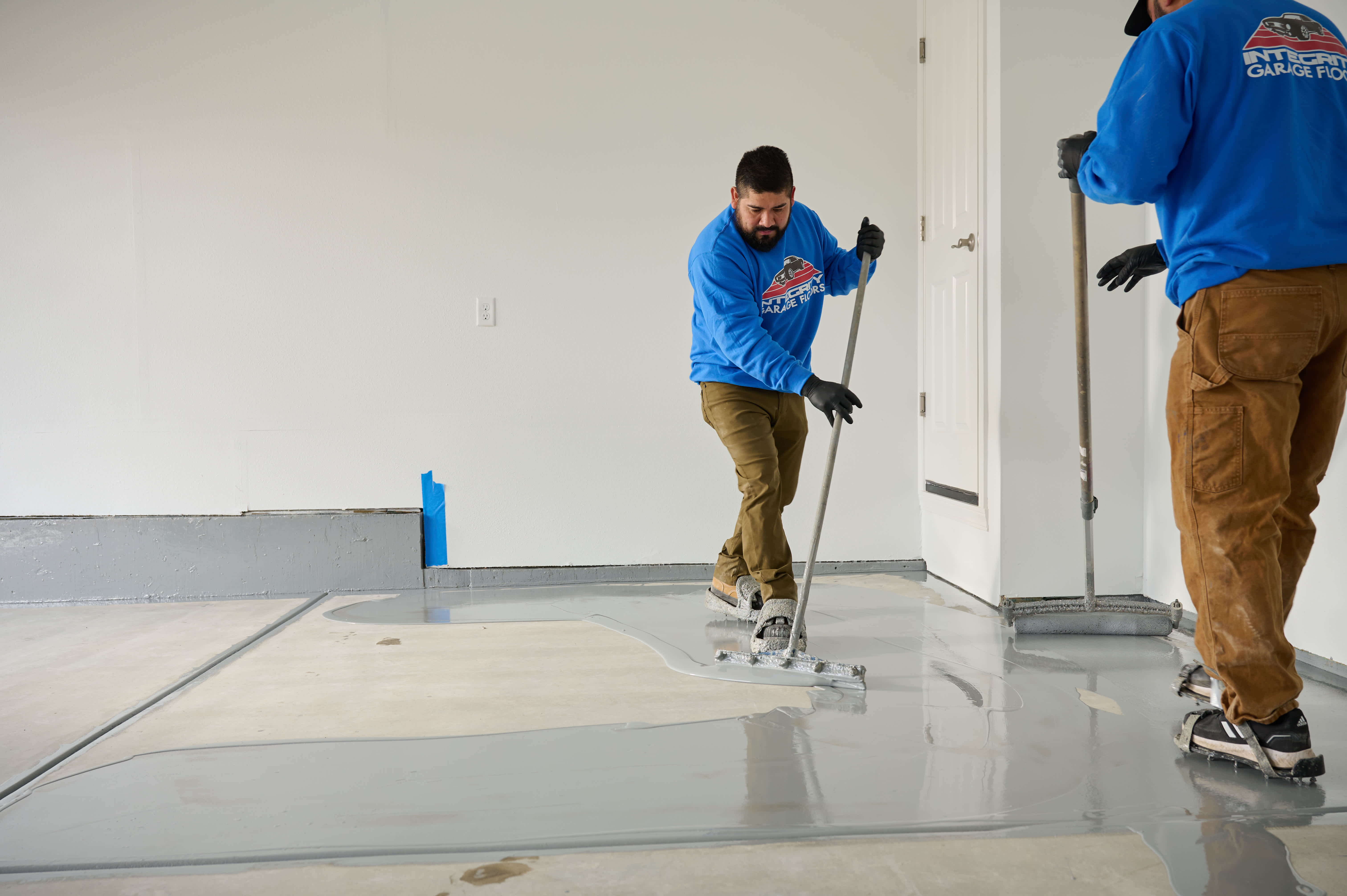 Two workers in blue shirts spreading gray epoxy coating on a garage floor using squeegees for a smooth, even finish.
