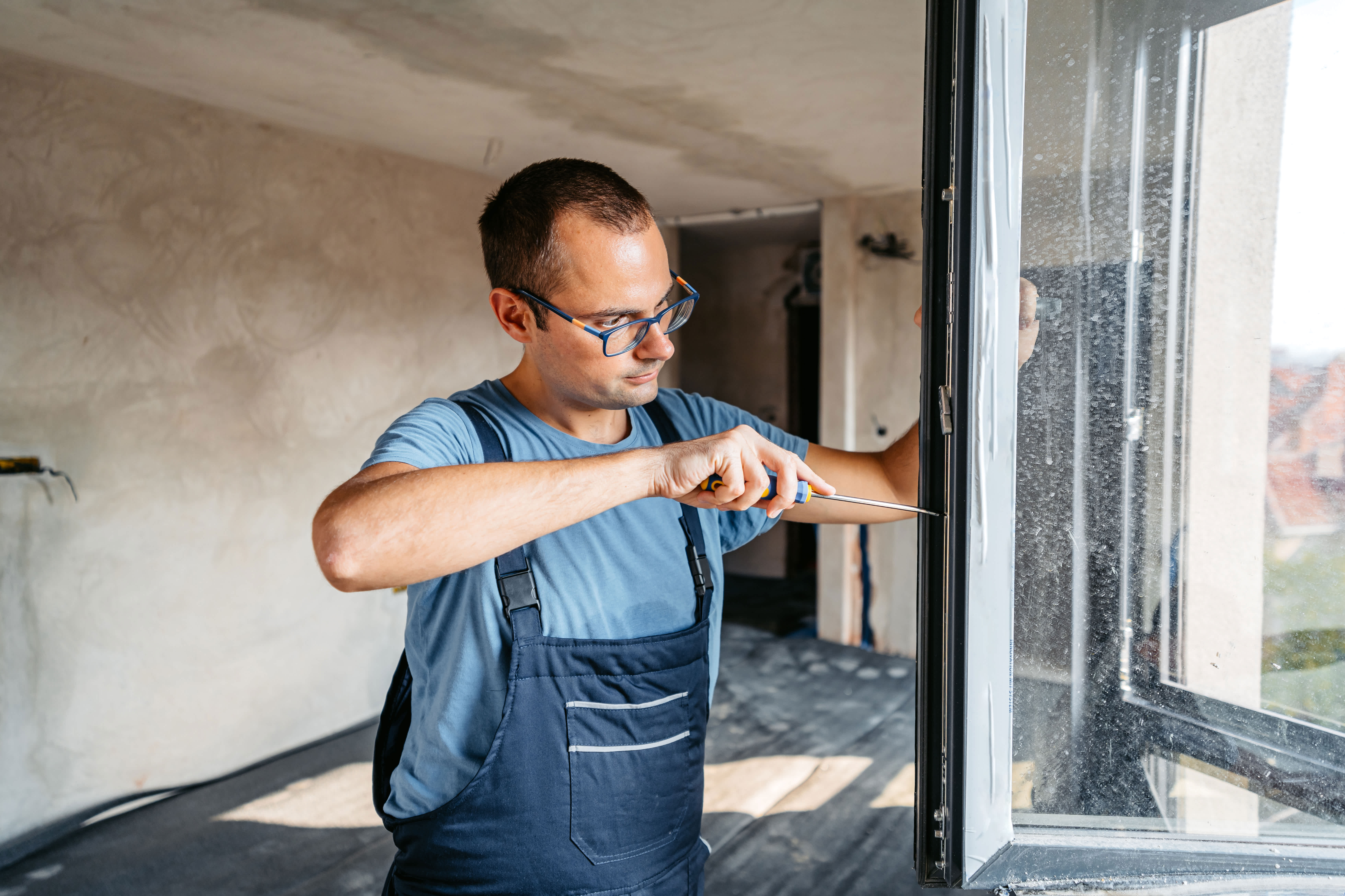 Repairman in overalls screwing the windows in an empty apartment under renovation.
