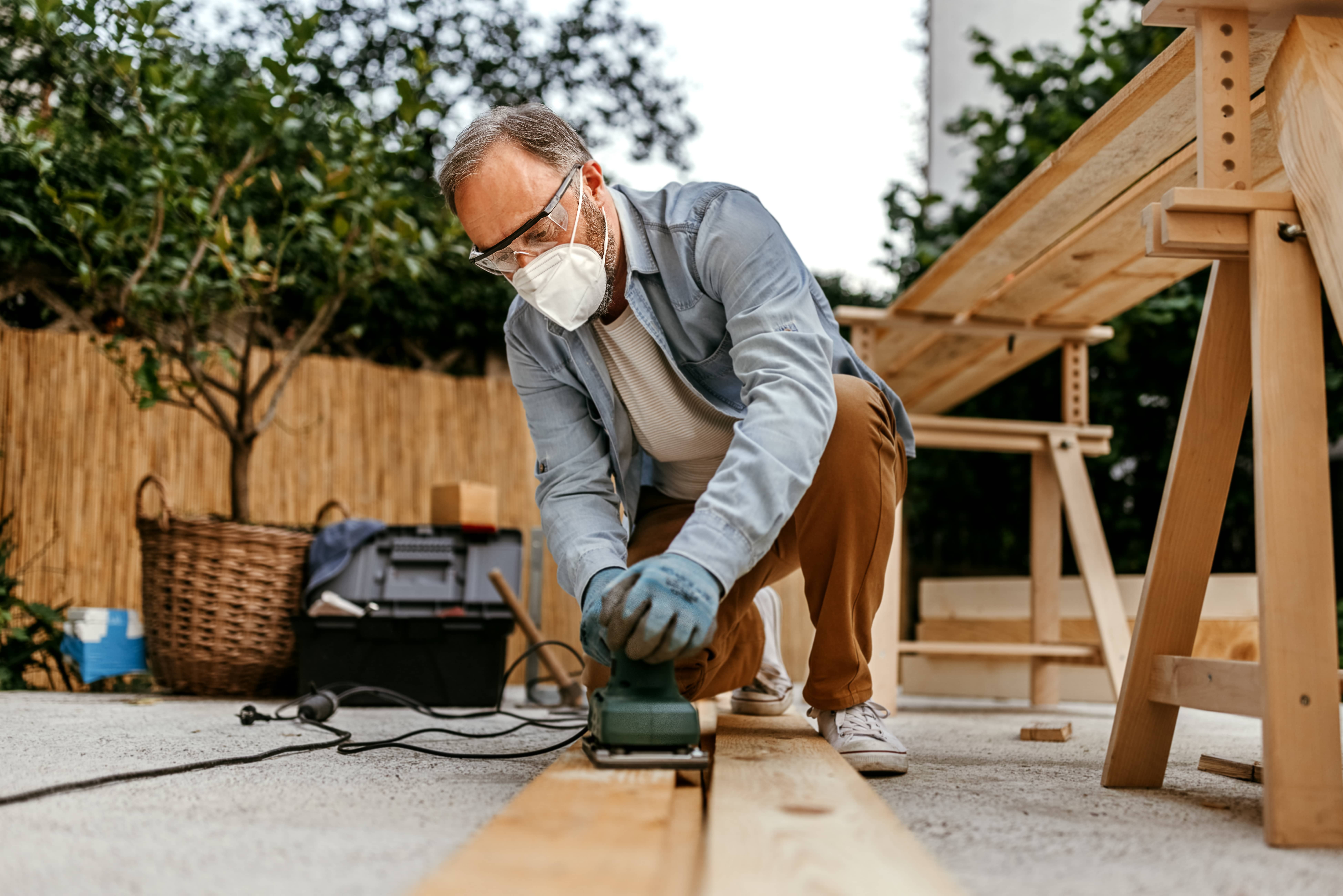 Man wearing a mask and safety goggles is sanding wooden furniture