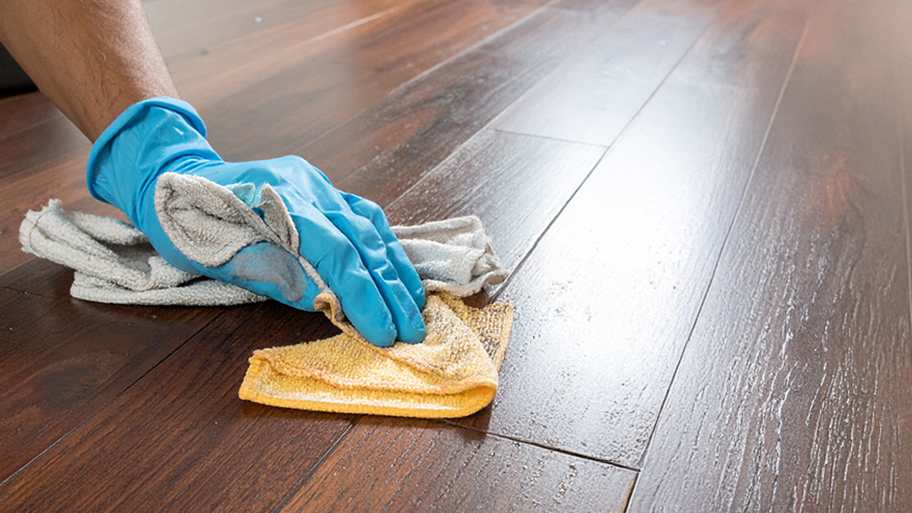 A hand in a blue glove wipes a glossy, dark wood floor with a yellow cloth