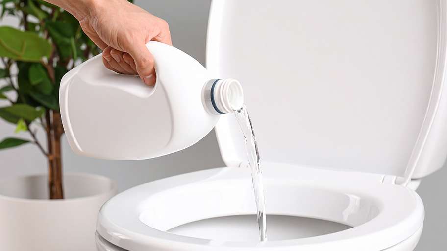 A hand is shown pouring a clear liquid, possibly bleach, from a white plastic container into a toilet bowl