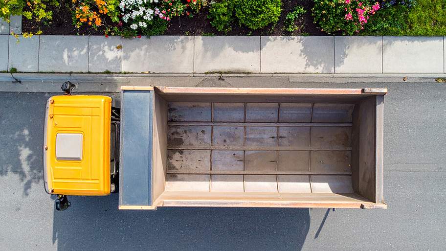 An aerial view of a bright yellow dump truck parked on an asphalt street with a sidewalk and planter box