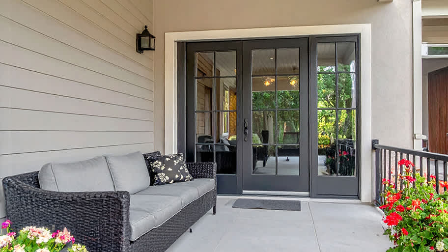 A covered patio features black-framed French doors, a gray wicker sofa with cushions, and potted red flowers.