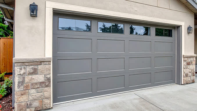 A light gray garage door with windows is set into a tan stucco house with stone accents and a concrete driveway.