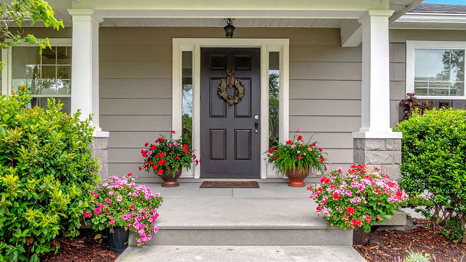 A symmetrical front porch features a dark brown door with a wreath, flanked by glass panels, white columns, and vibrant red and pink flowers.