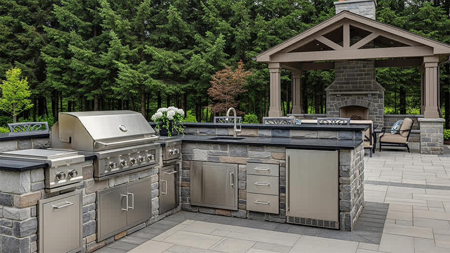 A spacious outdoor kitchen with a stone-clad grill and sink, a stainless steel refrigerator, and a covered patio area with a fireplace.