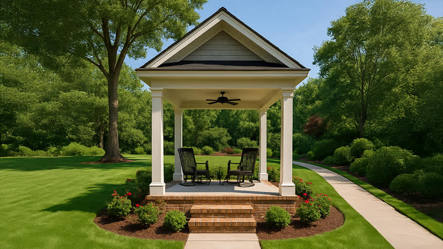 Backyard pavilion with a ceiling fan, stone floor, and seating, framed by lush green lawn and trees