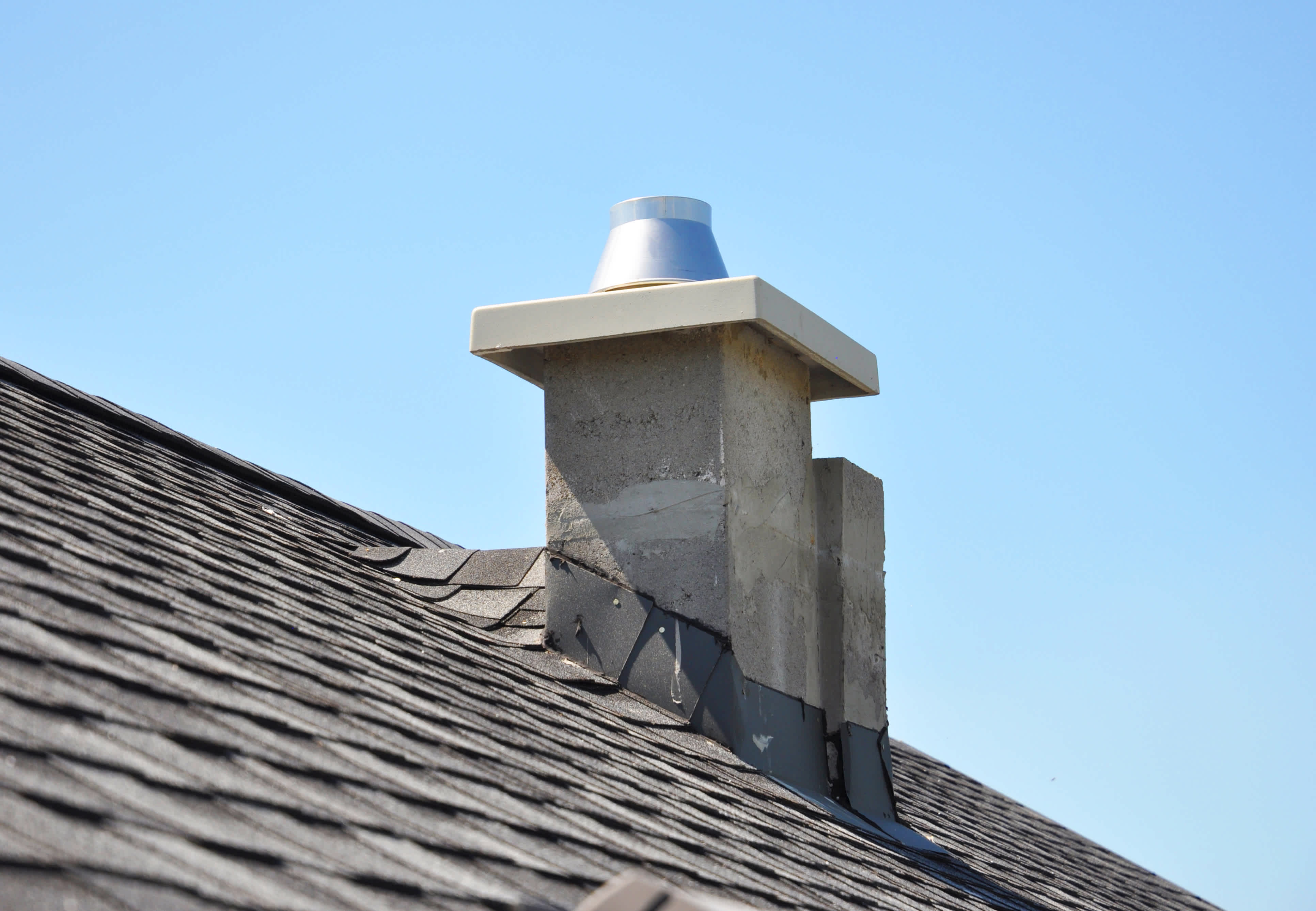 Brick chimney with metal cap and flashing on a shingled roof with a roof cricket behind it.