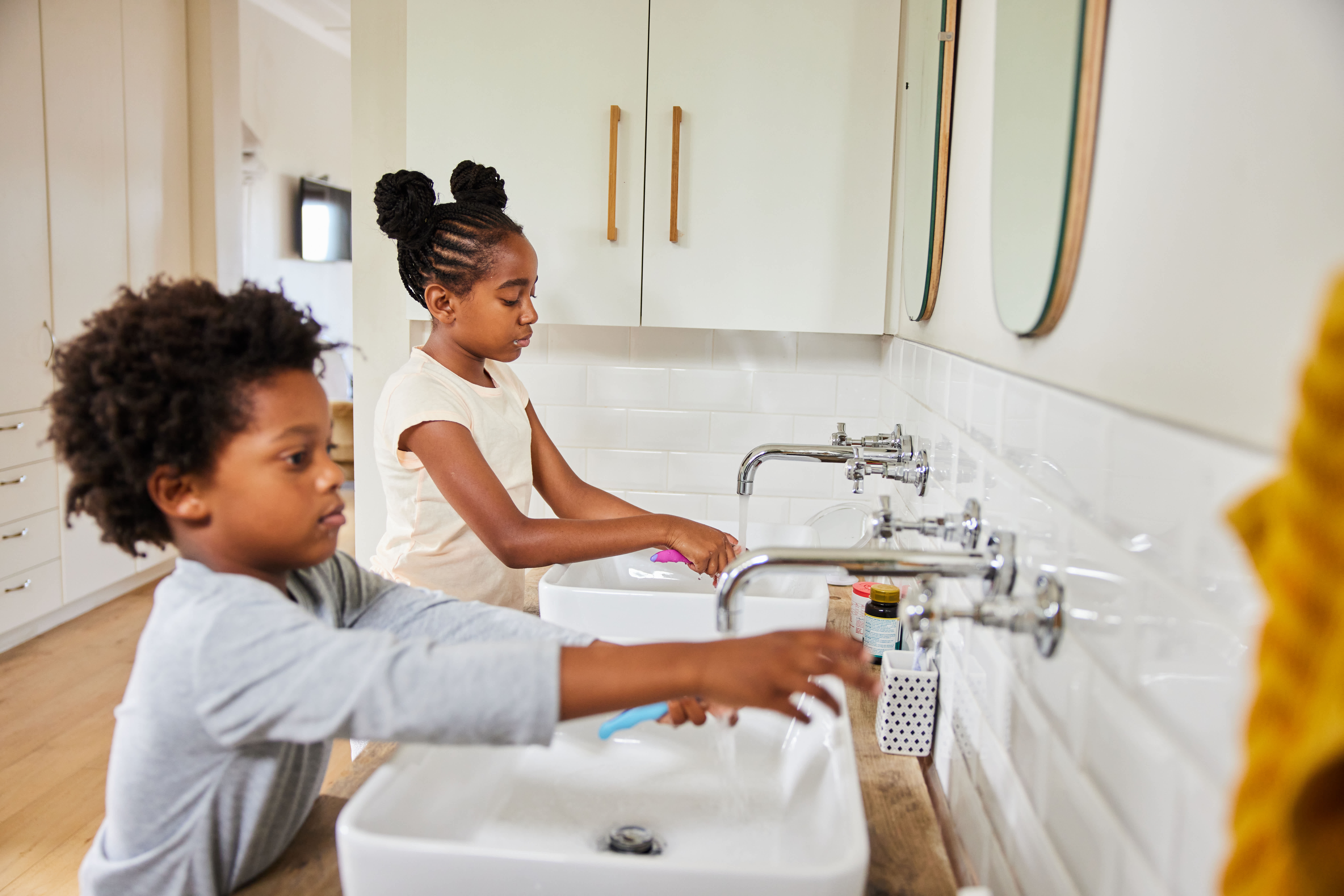 Two children washing their hands at bathroom sinks with toothbrushes nearby.