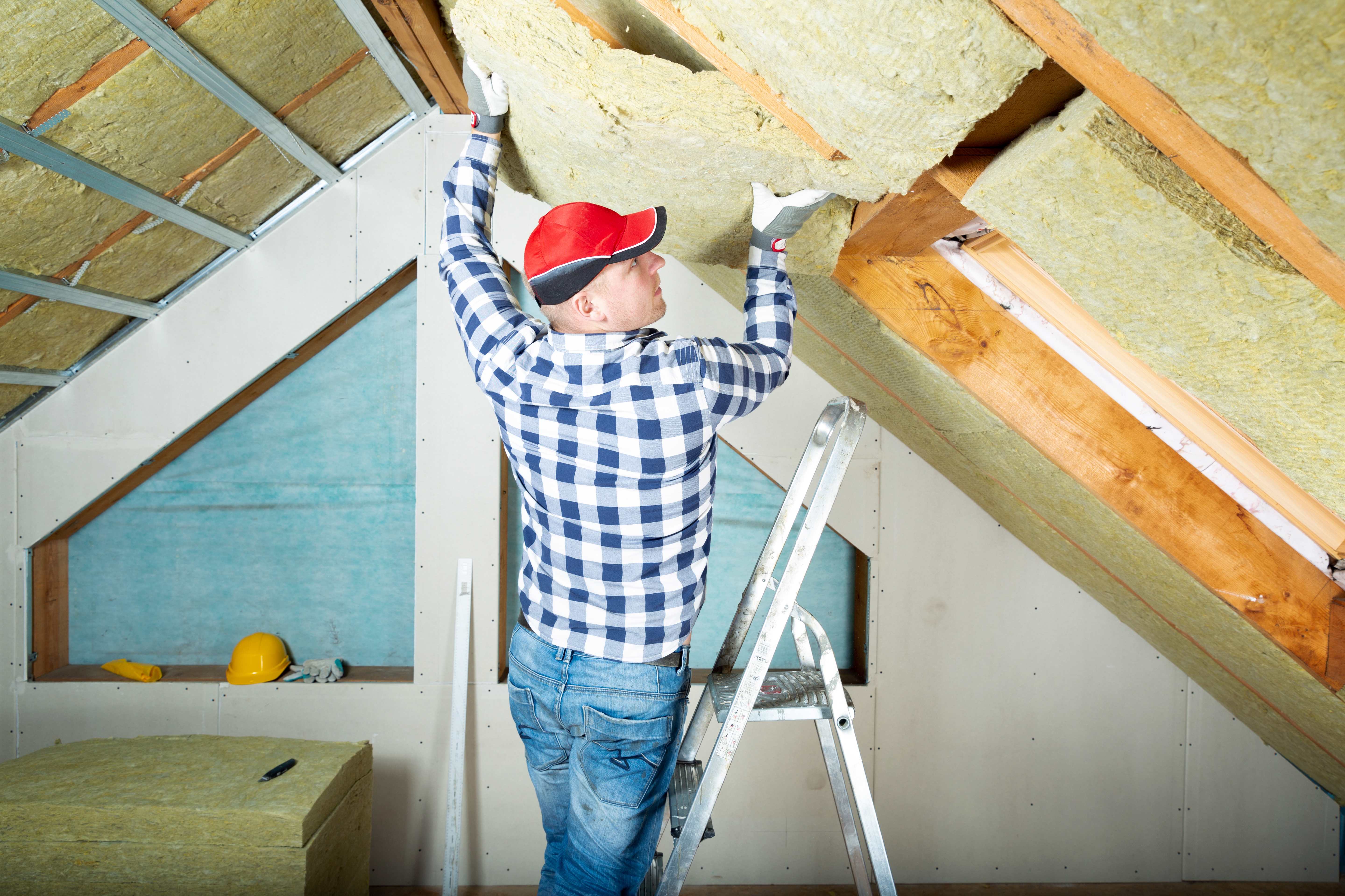 Worker installing mineral wool insulation in an attic ceiling while standing on a ladder