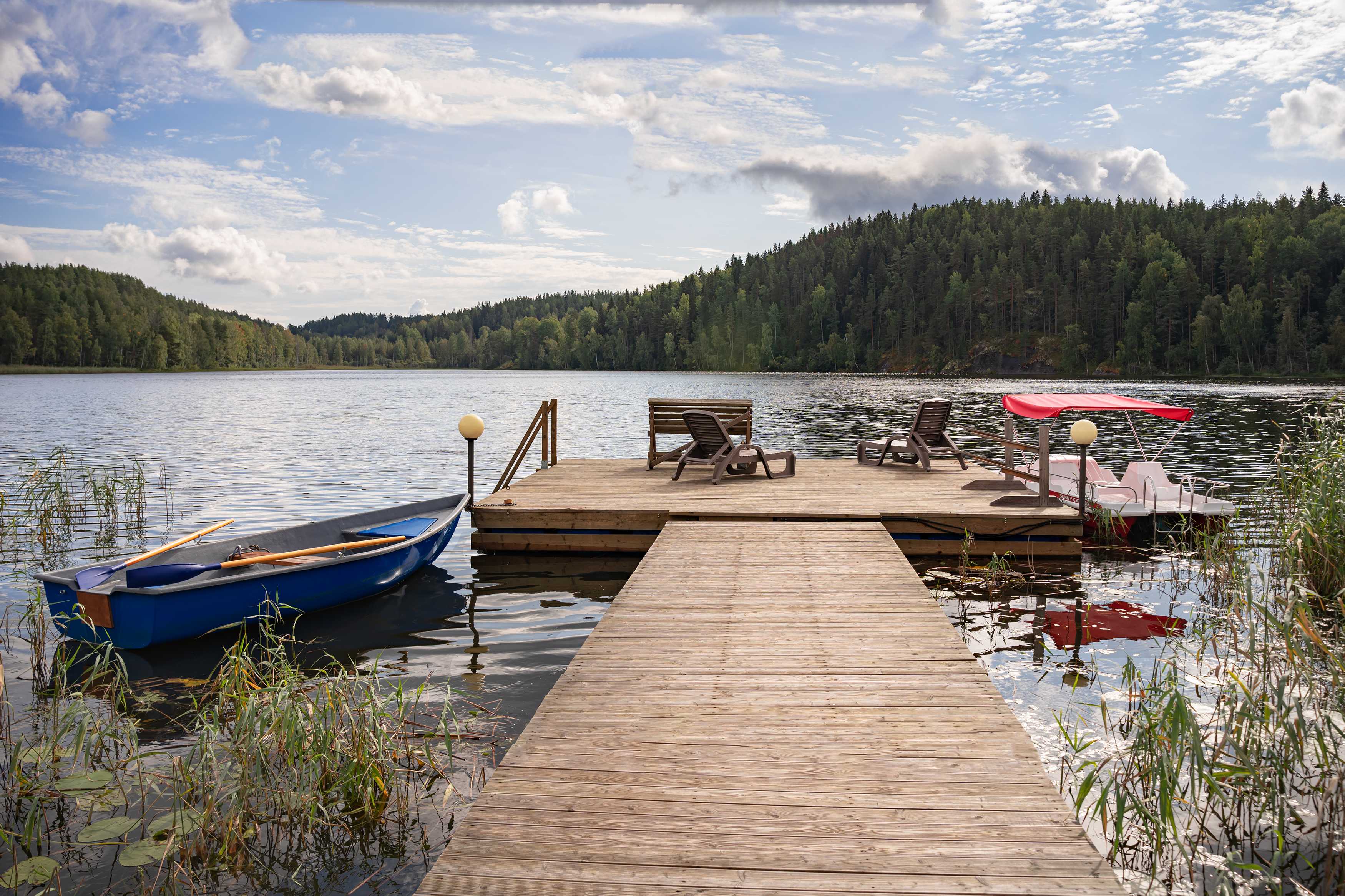 Wooden dock with lounge chairs, rowboat, and pedal boat on a calm forest lake.