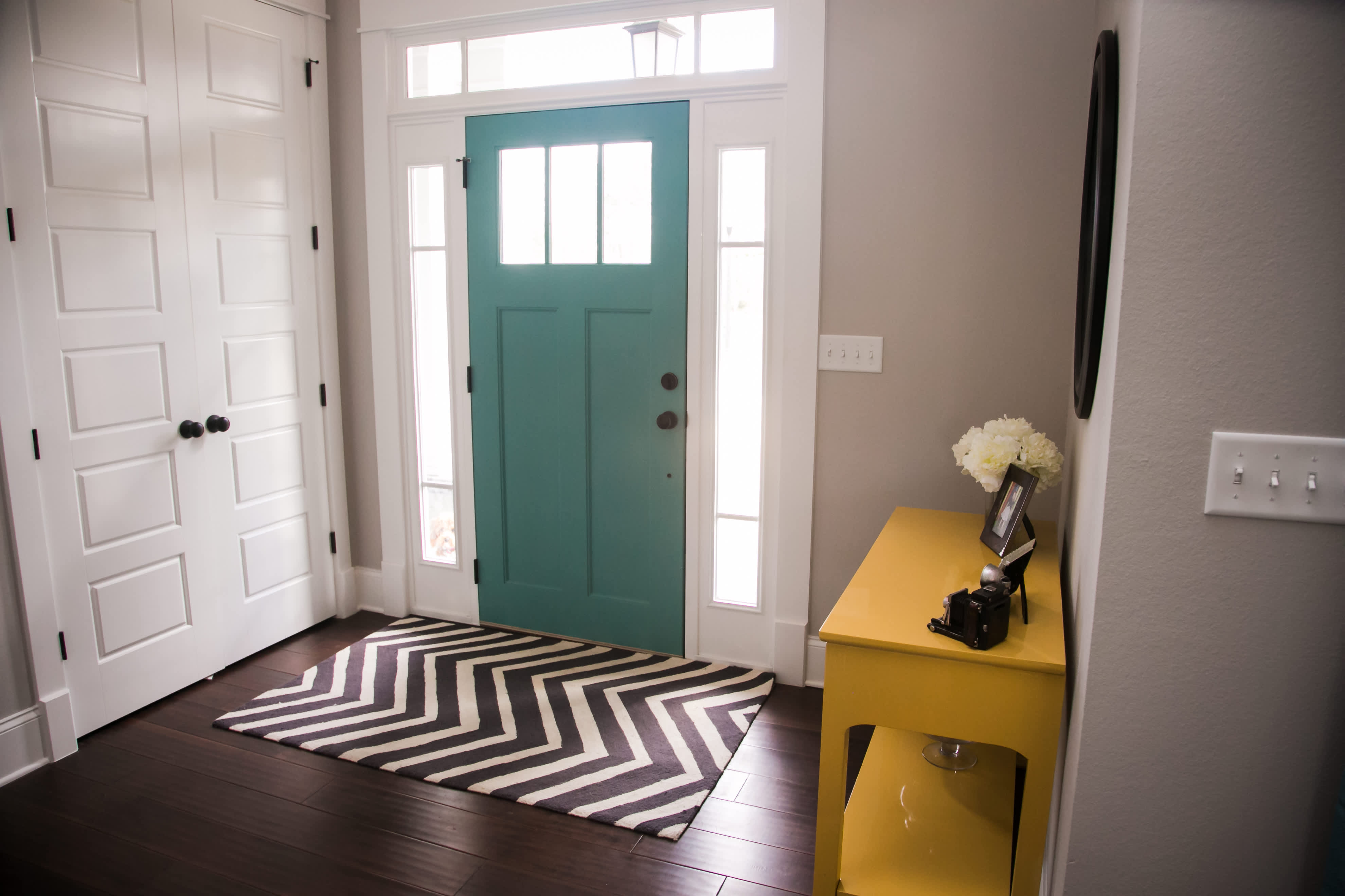 Bright entryway with a teal front door, white trim, chevron rug, and yellow accent table.