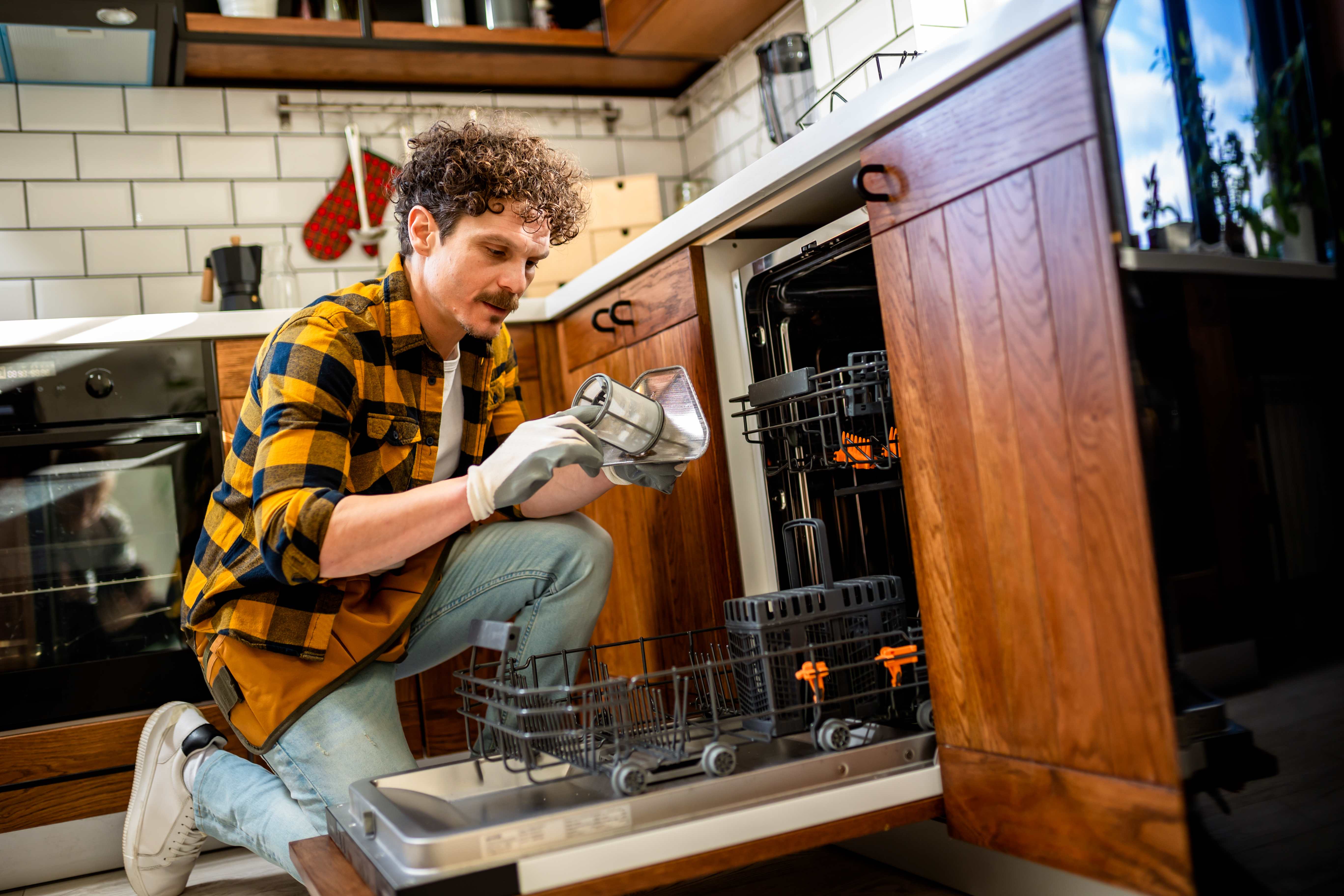 Man wearing gloves kneeling on the floor while cleaning the filter of a dishwasher in a kitchen