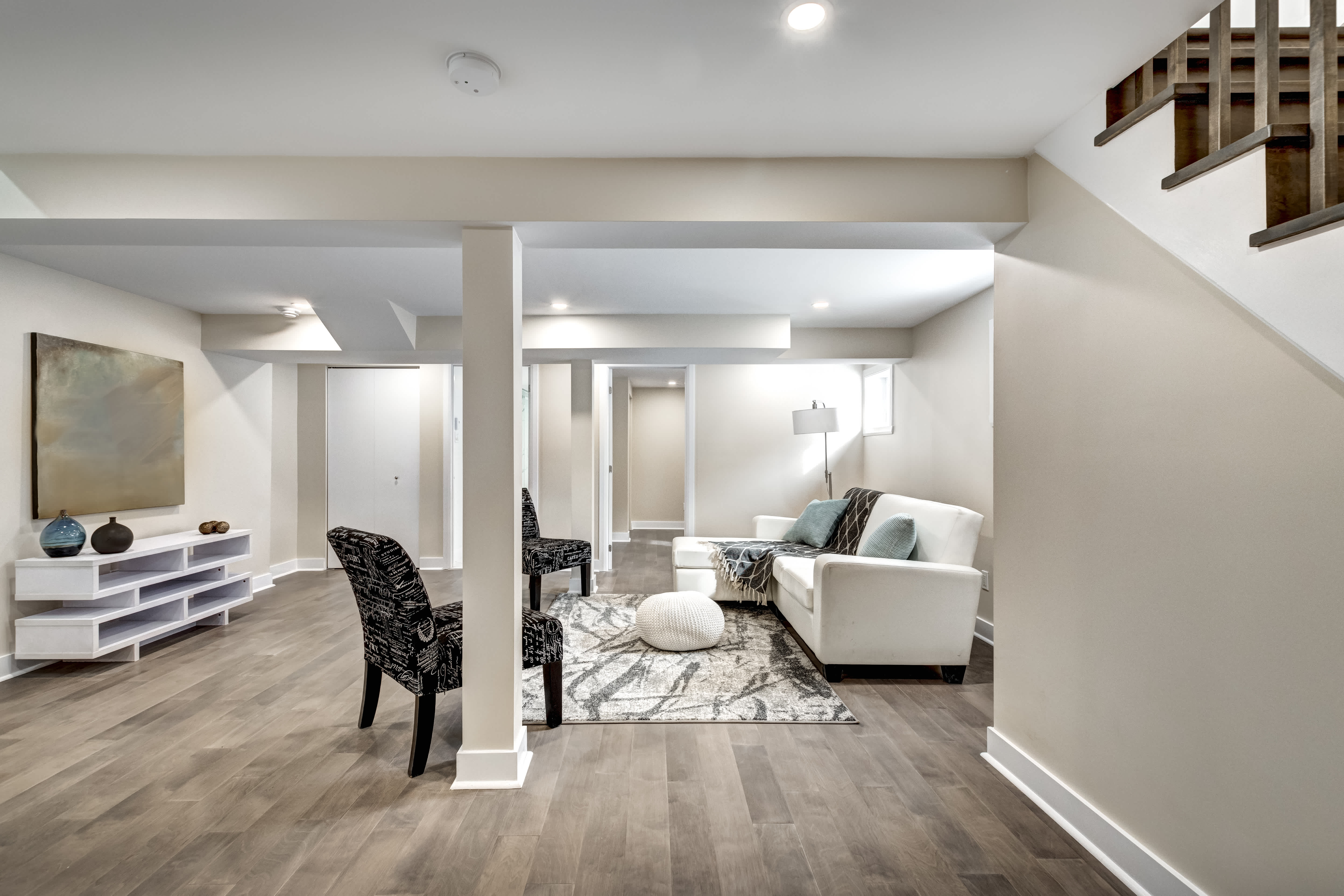 Finished basement with light wood flooring, white walls, and a furnished seating area including a white sofa, accent chairs, and a patterned rug
