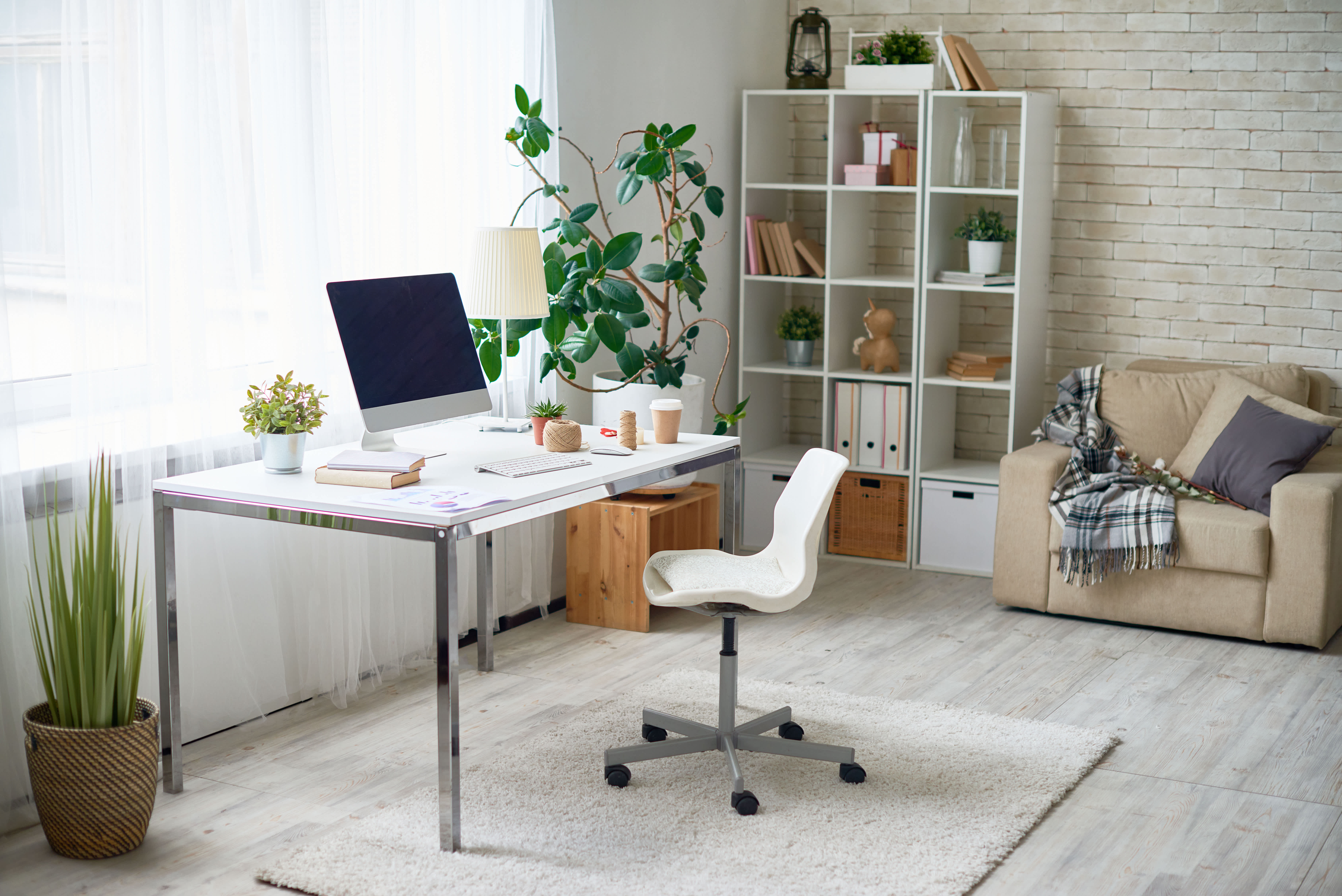 Bright home office with computer on white desk, indoor plants, bookshelves, and cozy armchair with blanket